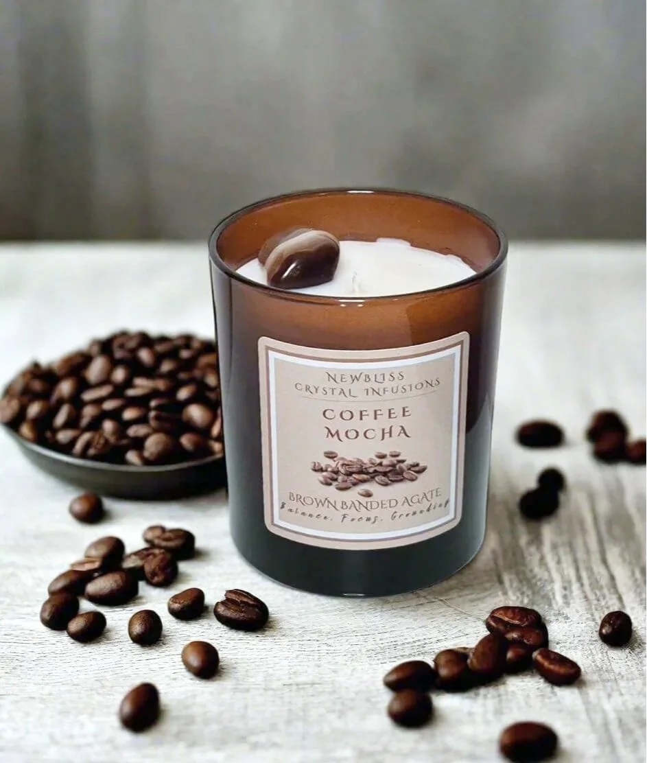 A brown glass candle labeled 'Coffee Mocha' surrounded by roasted coffee beans and a bowl of more coffee beans on a white wooden surface.
