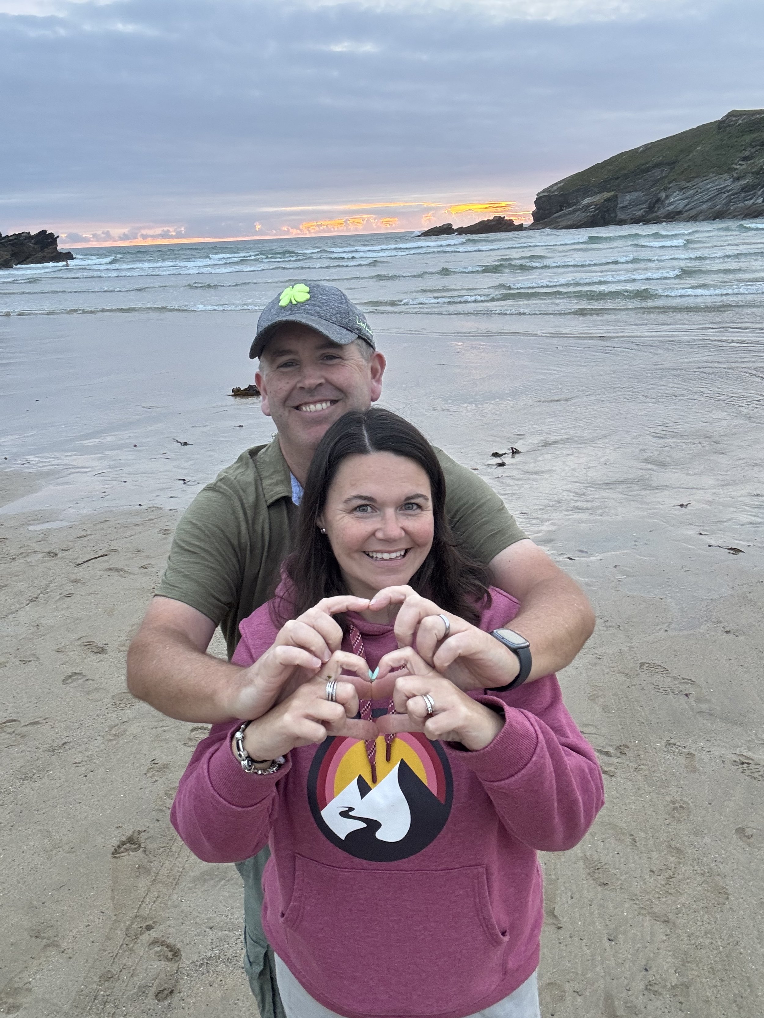 A smiling couple on a beach at sunset, making a heart shape with their hands.