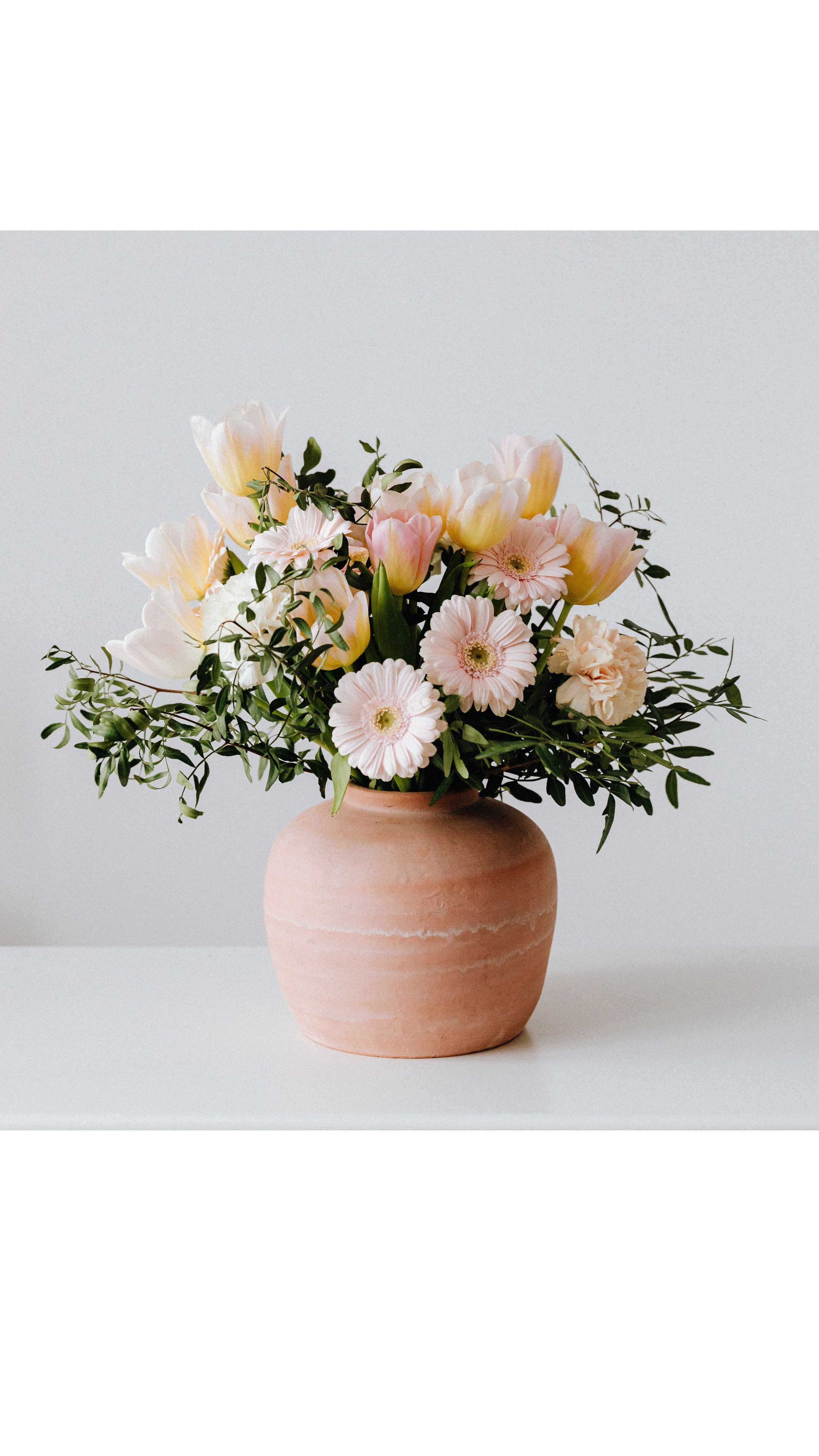 A pink ceramic vase filled with light pink and white tulips, daisies, and greenery on a white surface against a plain white background.