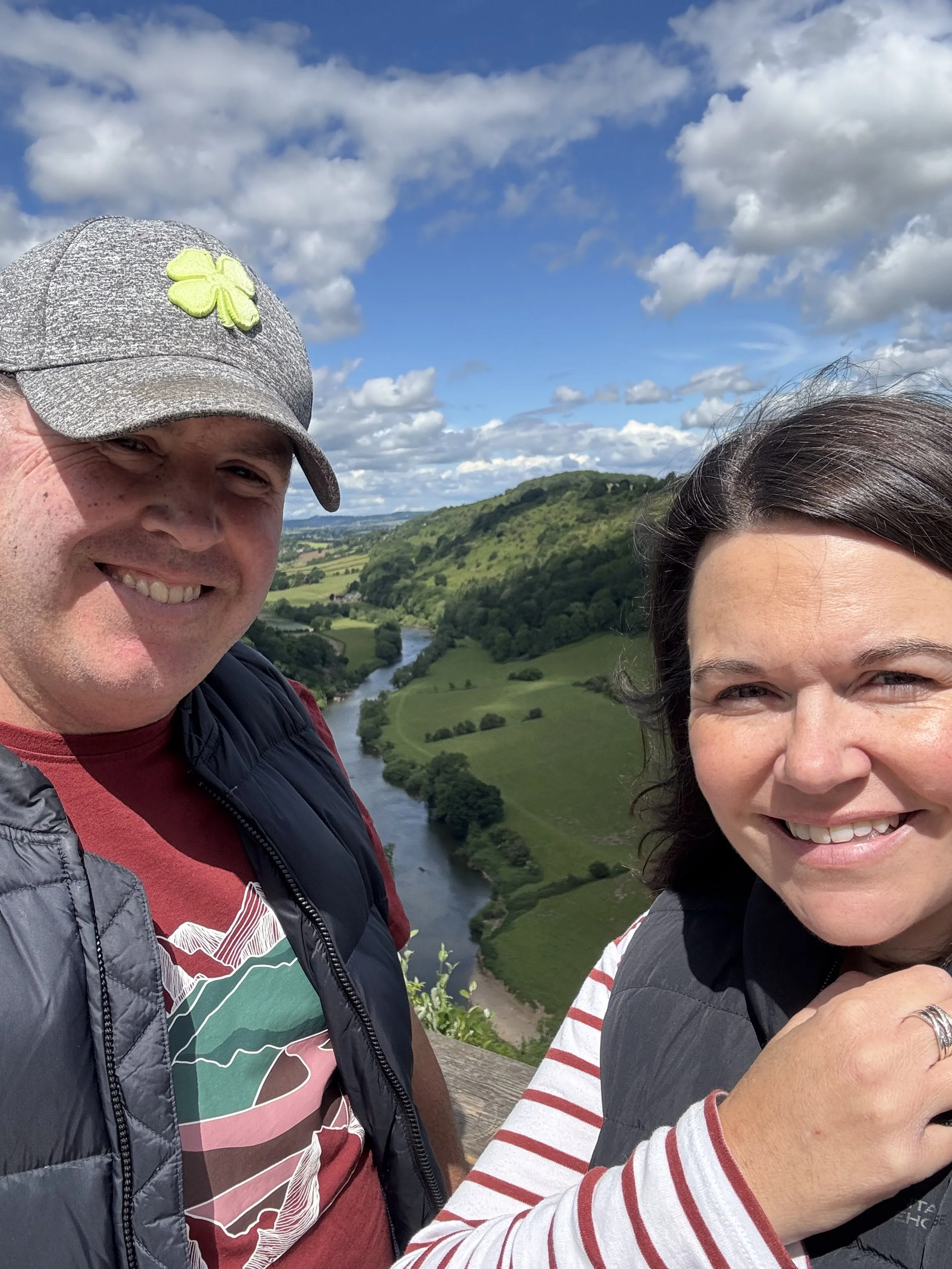 Smiling man and woman taking a selfie outdoors with a scenic view of a river, green hills, and partly cloudy sky in the background.