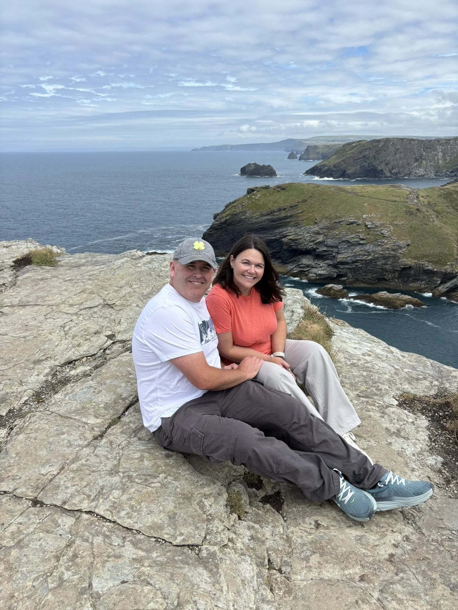 Lisa & Chris sitting on a rocky cliff overlooking the ocean, with green cliffs and rock formations in the background.