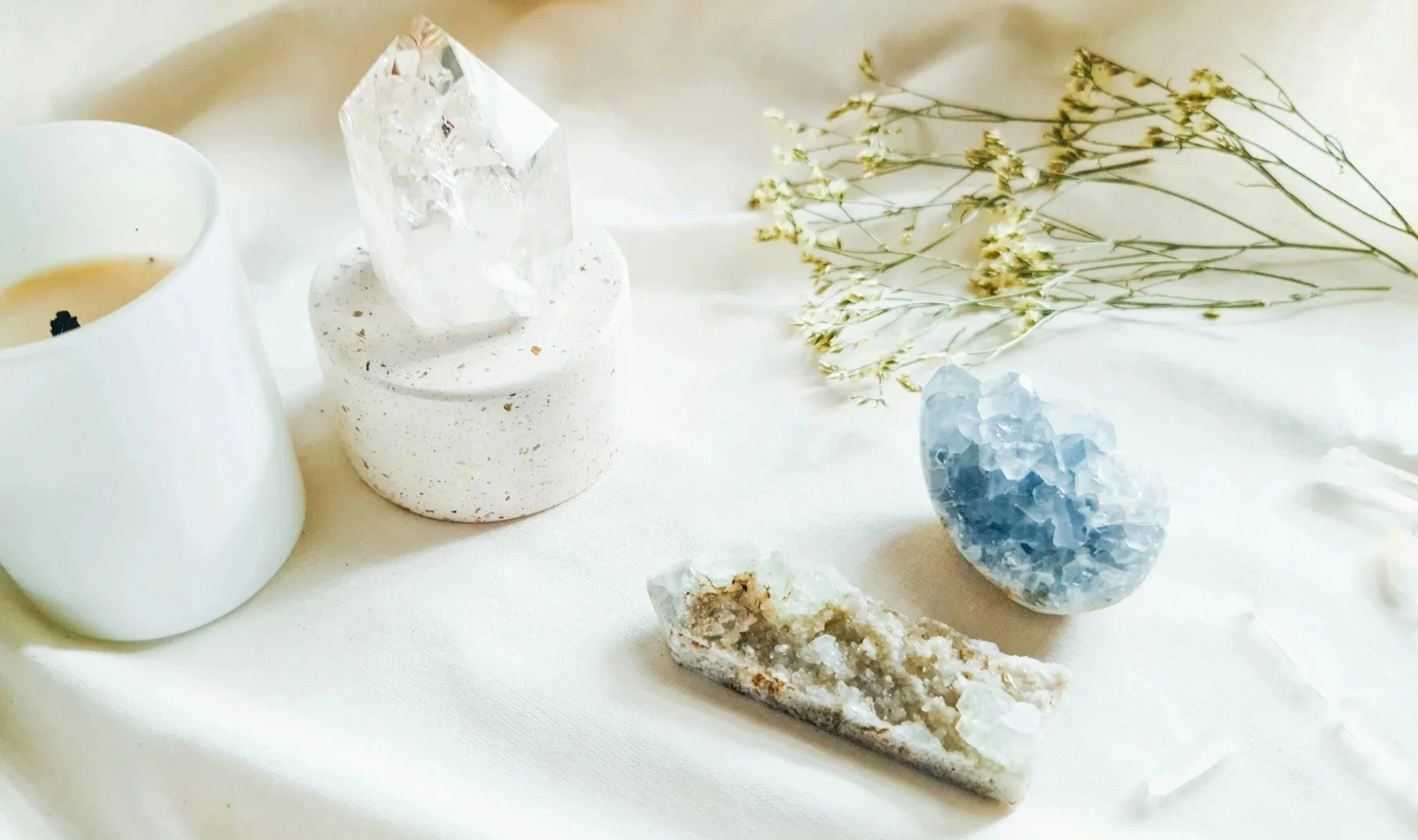 A white candle with a black wick, a clear quartz crystal on a white speckled holder, a dried flower branch, a blue geode, and a stone with white and beige crystals on a white surface.