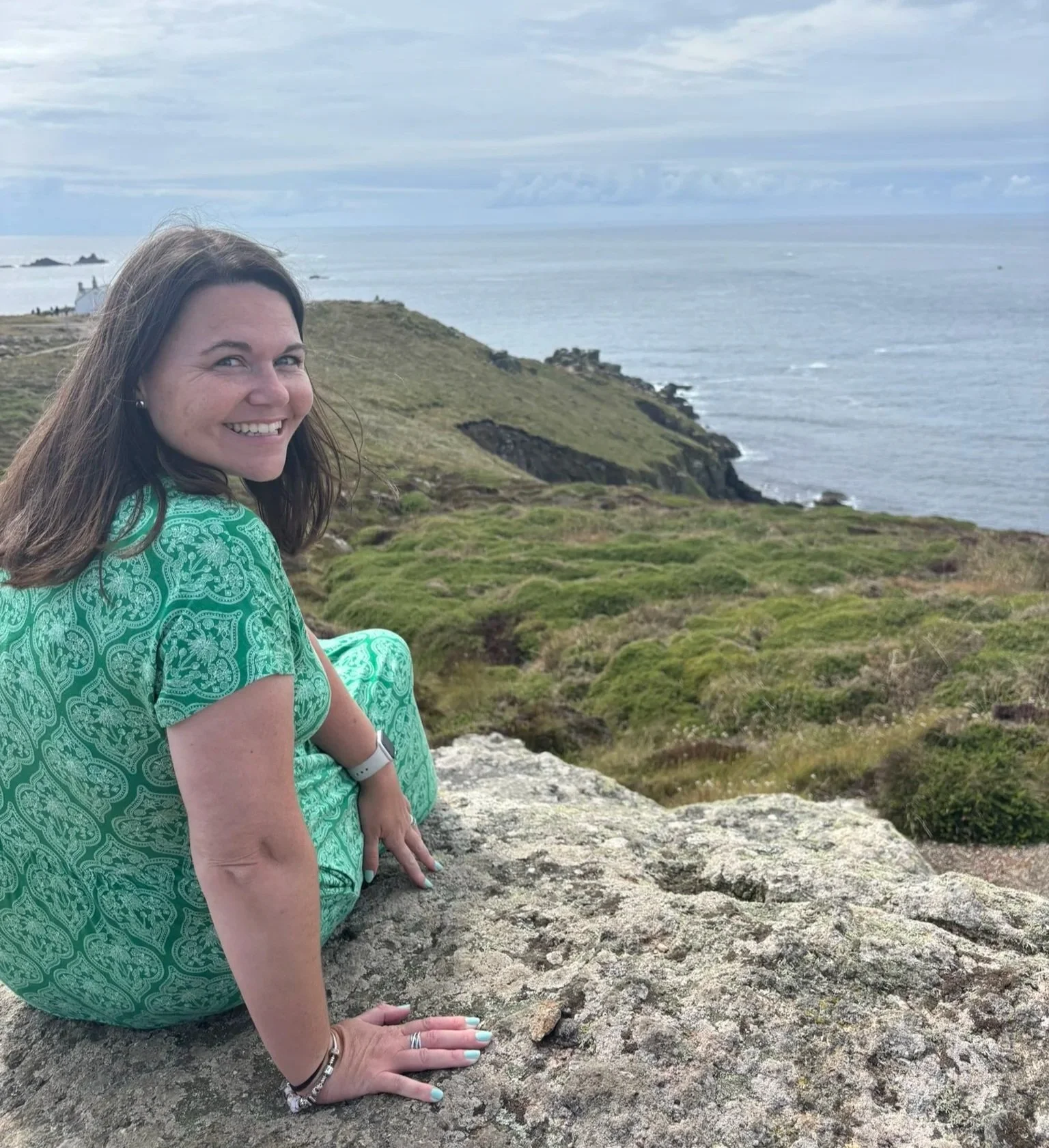 A woman with long brown hair sitting on a large rock, smiling at the camera with a scenic coastal landscape in the background consisting of green hills, ocean, and cloudy sky.