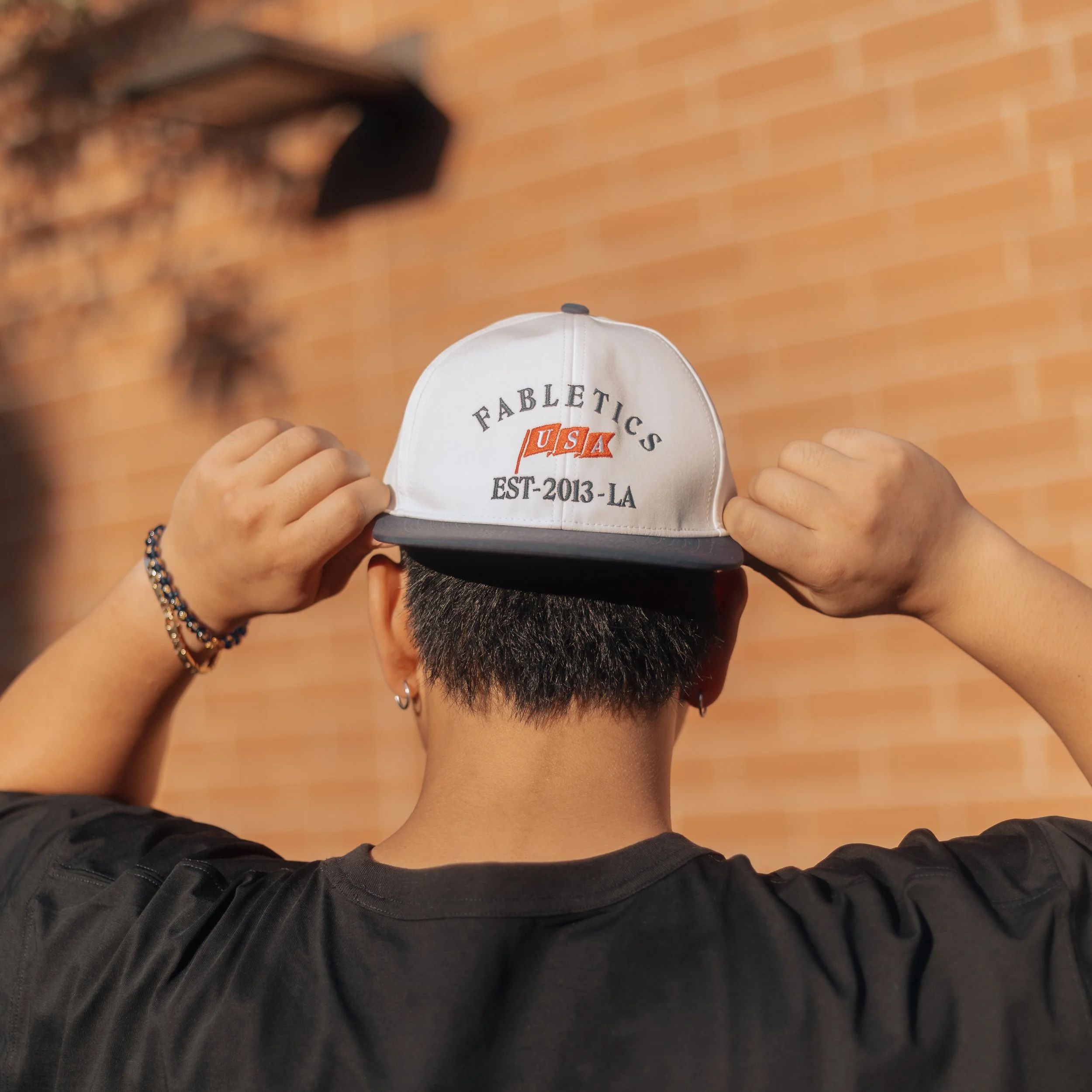 A person is holding up a white cap with embroidered text and logo on the front. The background shows a wooden floor.
