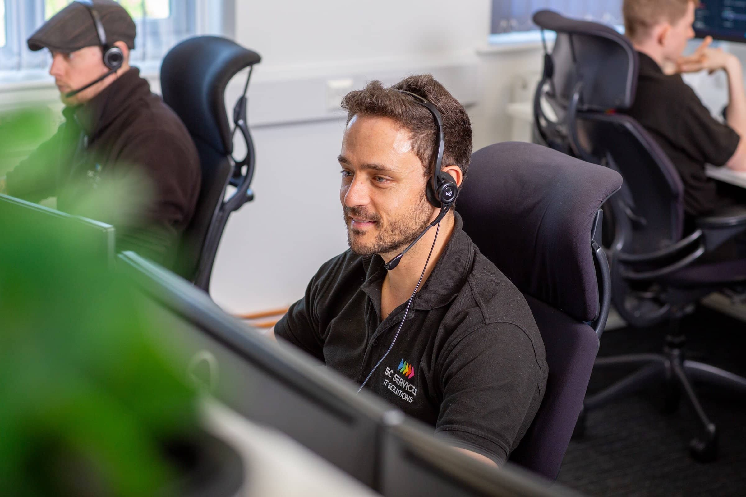 Man wearing a black polo shirt with a logo, sitting at a computer desk in an office, wearing a headset, working on his computer.