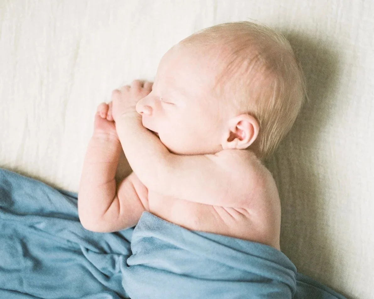 A sleeping baby with closed eyes, lying on a soft surface, with arms crossed and wrapped in a blue blanket.
