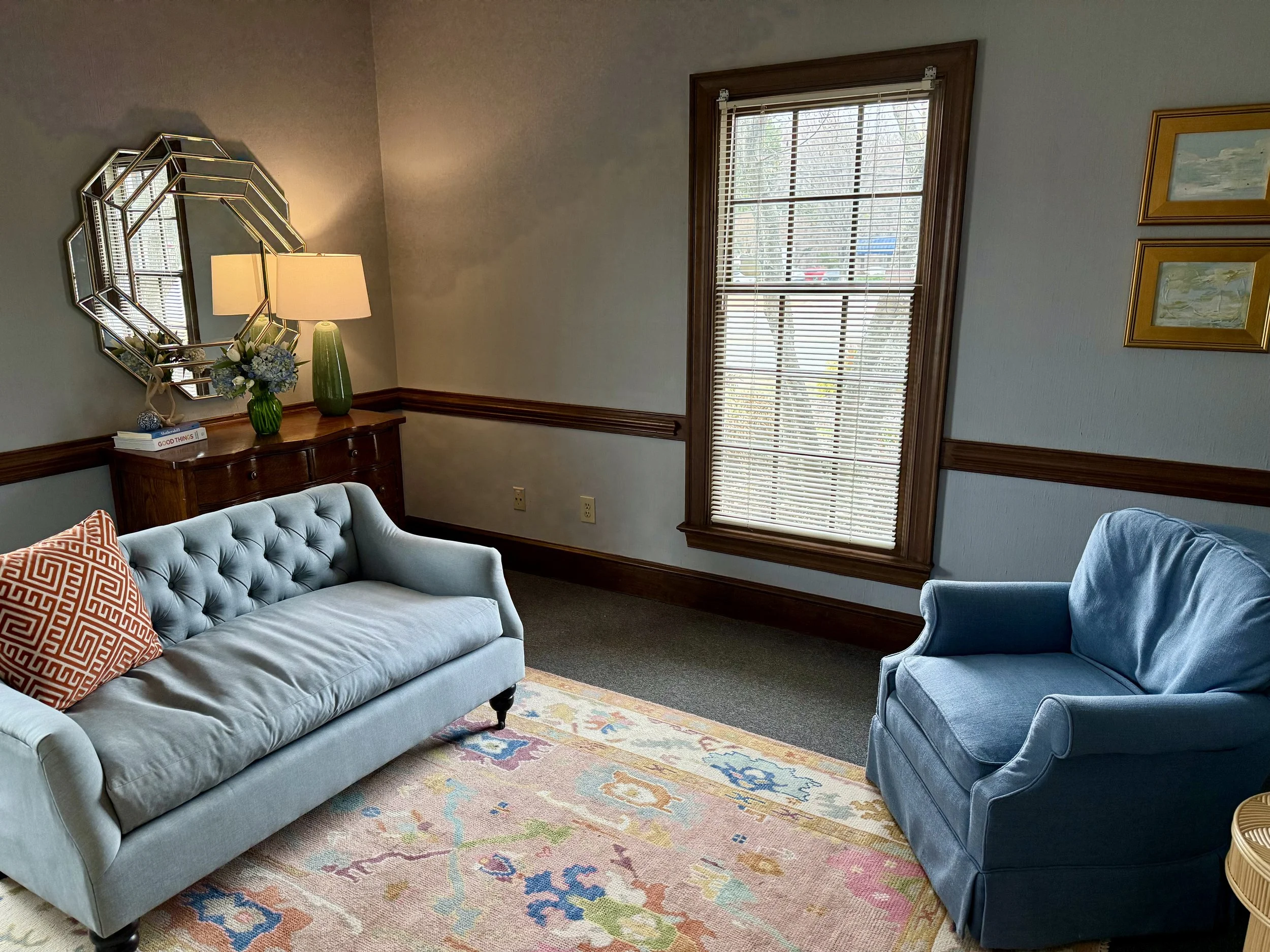 A cozy living room with a light blue tufted sofa on the left, an orange patterned pillow, a wooden sideboard with a green vase and flowers, and a cream-colored lamp. A blue armchair is on the right, and a colorful area rug covers the floor. The room has a large window with wooden framing and blinds, and wall art on the right side.