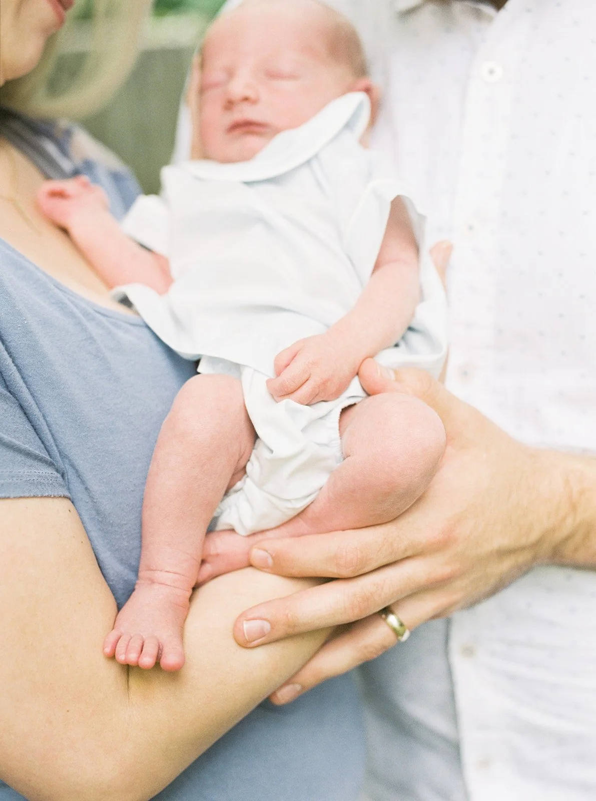 A newborn baby being held by an adult, wearing a white outfit, with the baby's eyes closed.