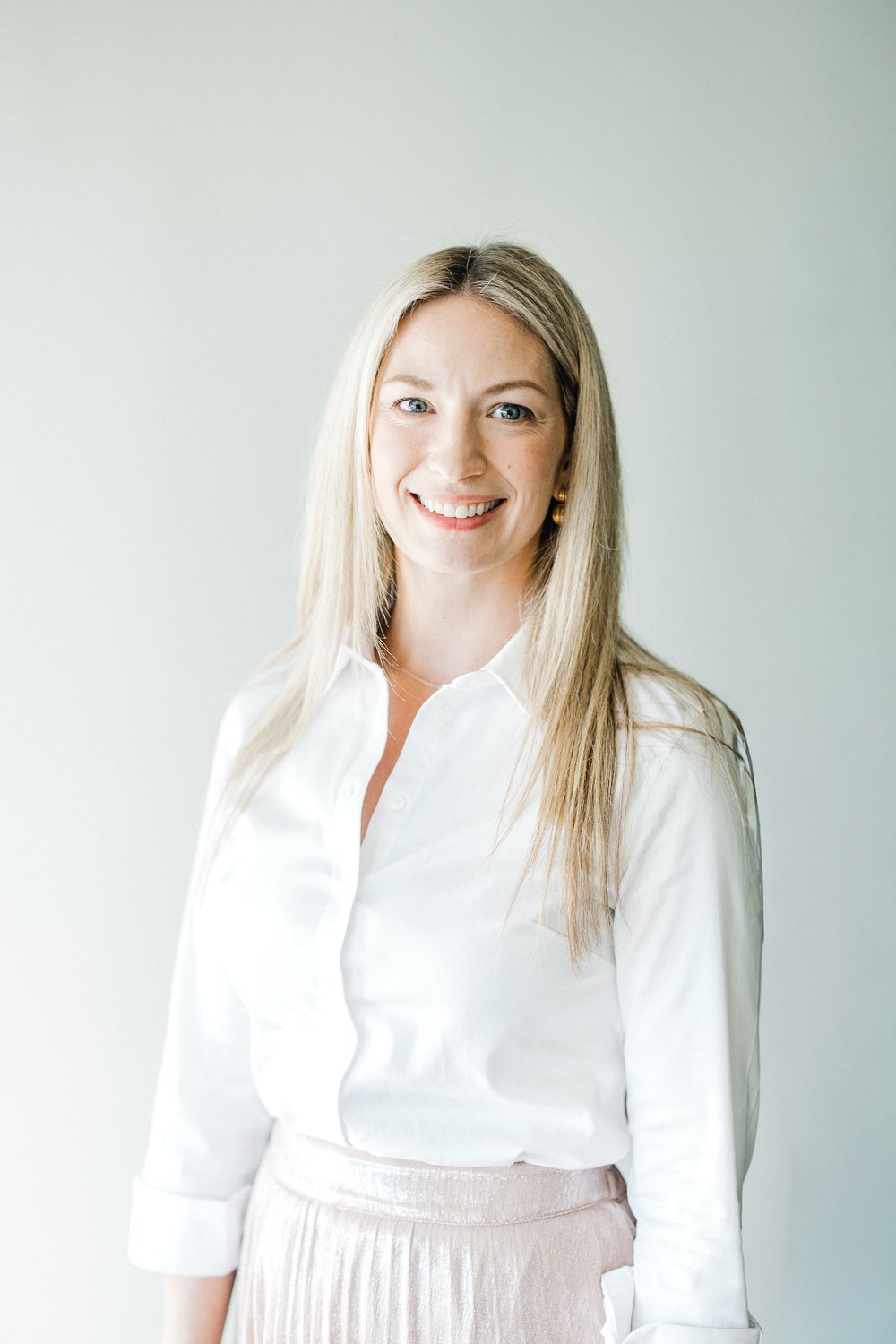 A smiling woman with blonde hair wearing a white blouse and light pink skirt, standing against a plain light-colored background.
