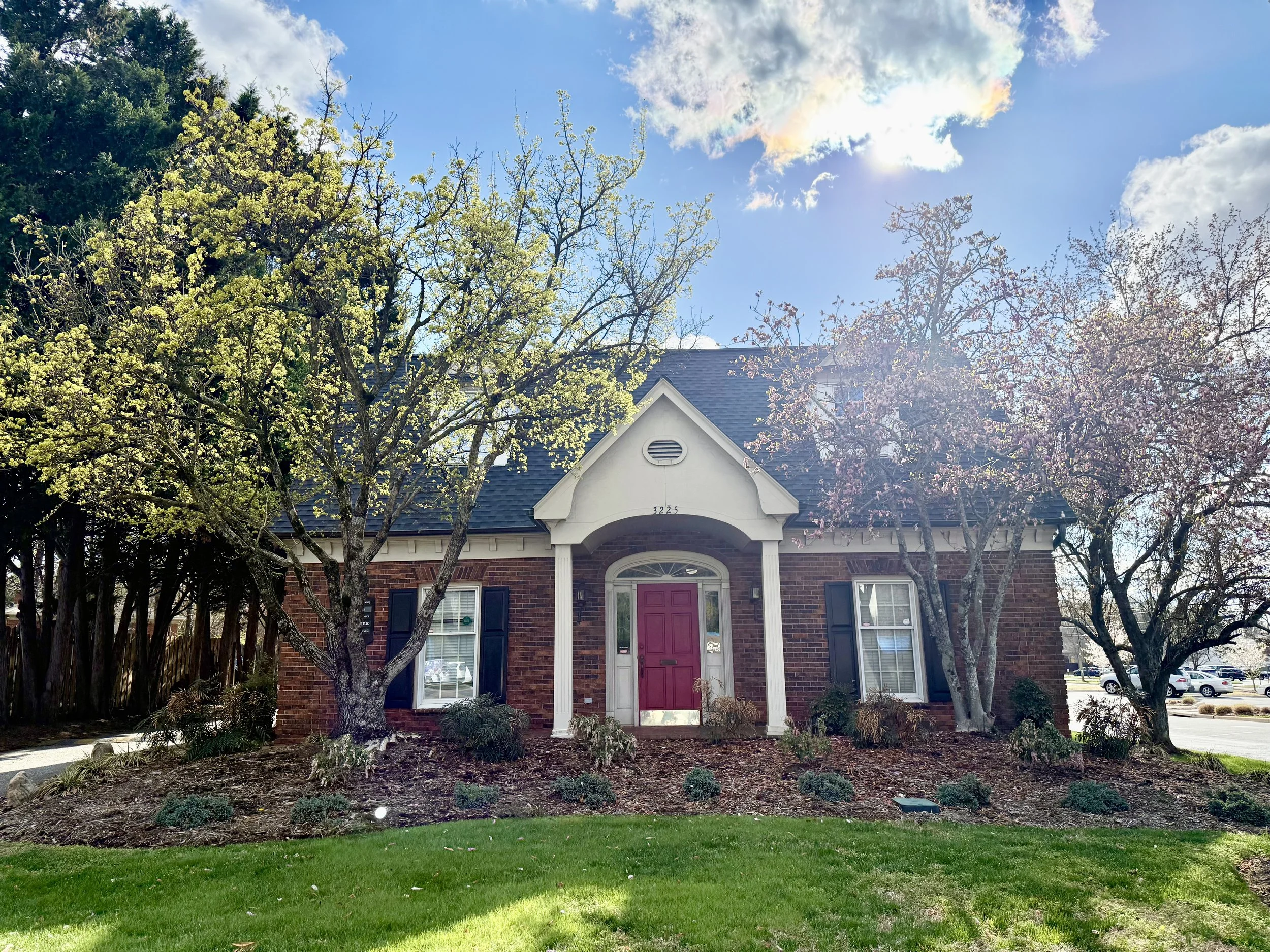 A brick house with a red front door, surrounded by trees with blossoming flowers, under a partly cloudy sky.
