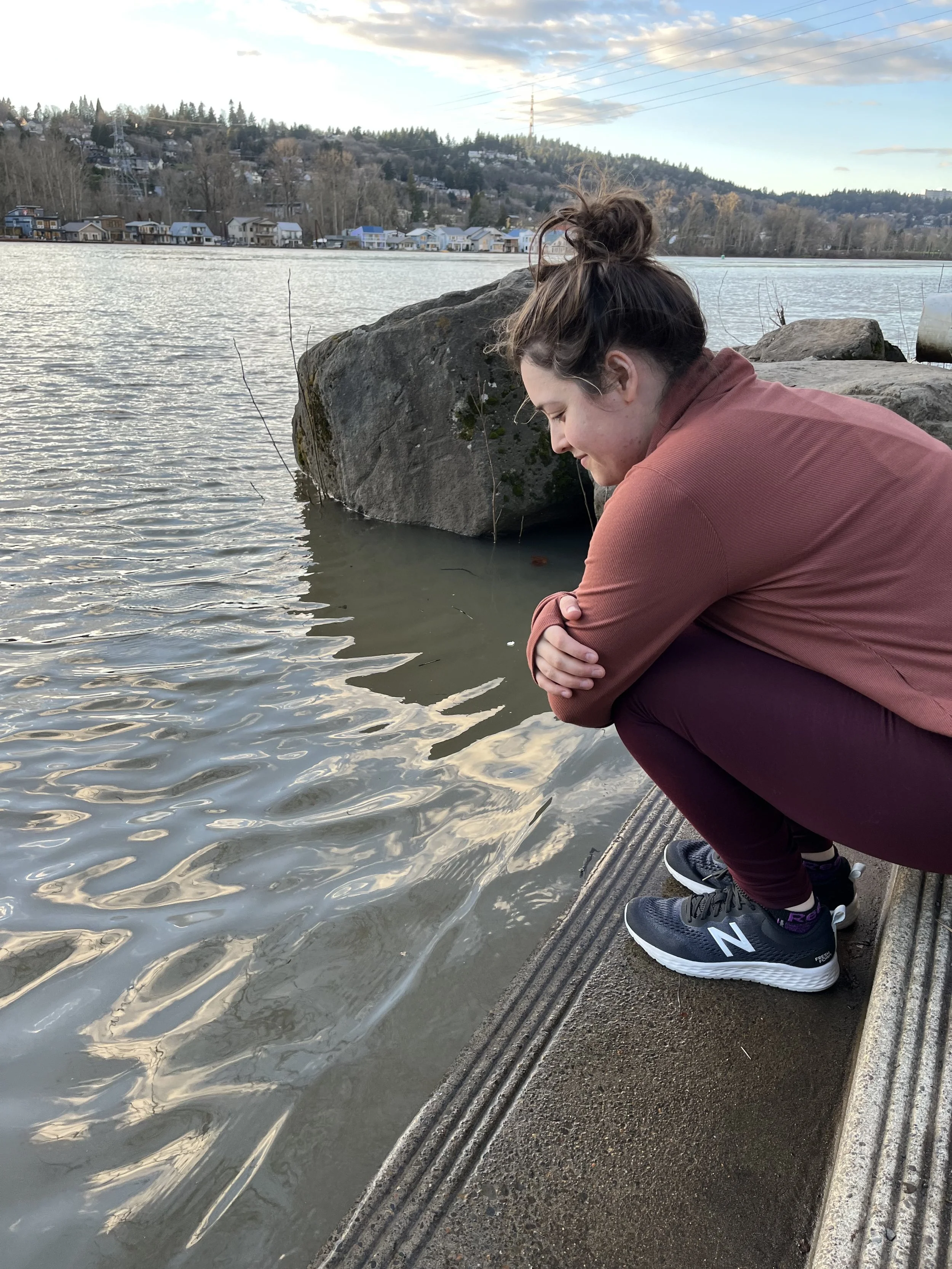 A woman crouching looking into a river and smiling, wearing black sneakers with white soles, brown pants, and a pinkish-brown jacket, with a few large rocks in the background