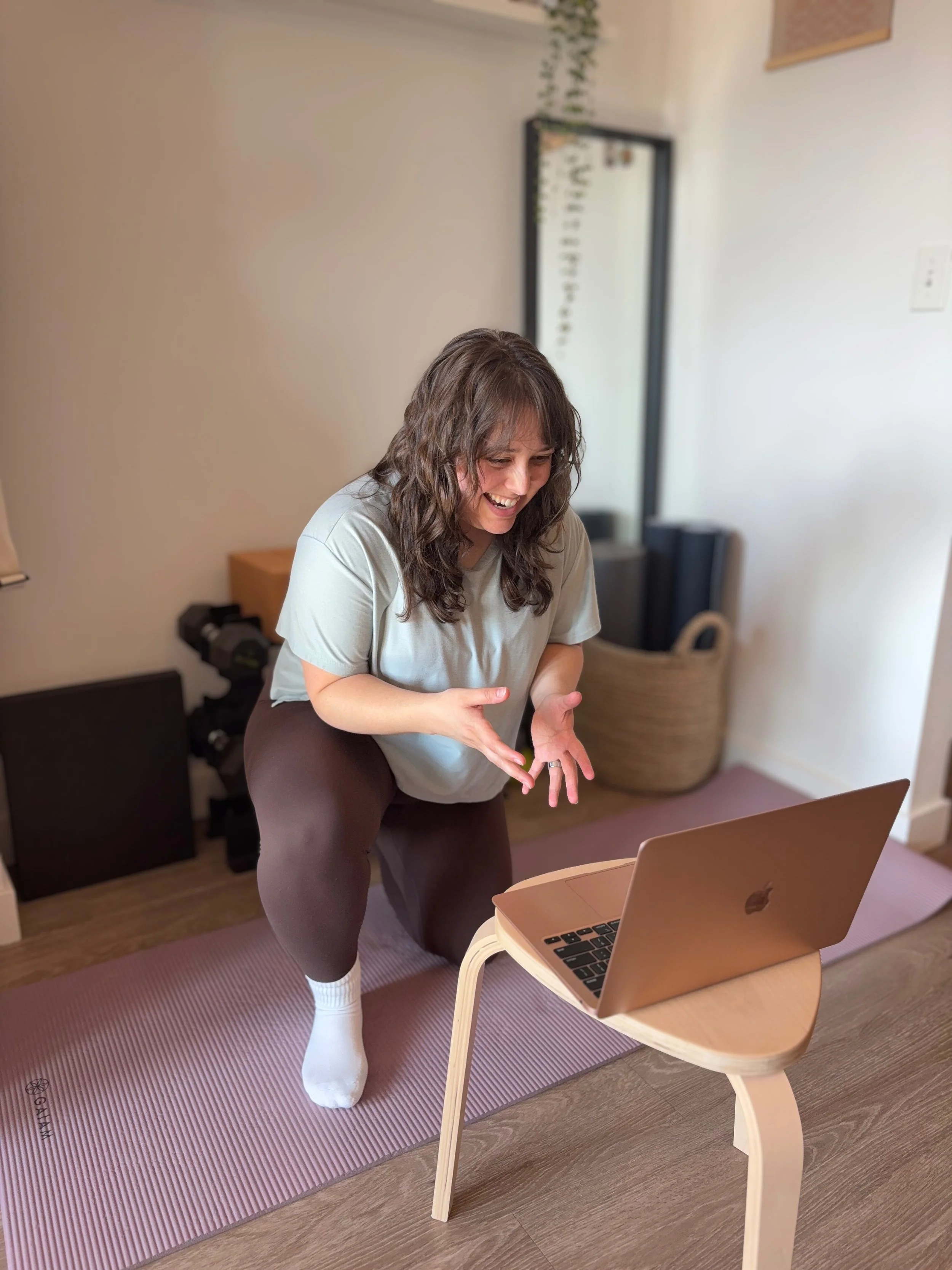 A woman smiling while kneeling on a pink yoga mat in front of home exercise equipment, with a laptop on a small wooden table in front of her. She is interacting with an unseen client on the screen
