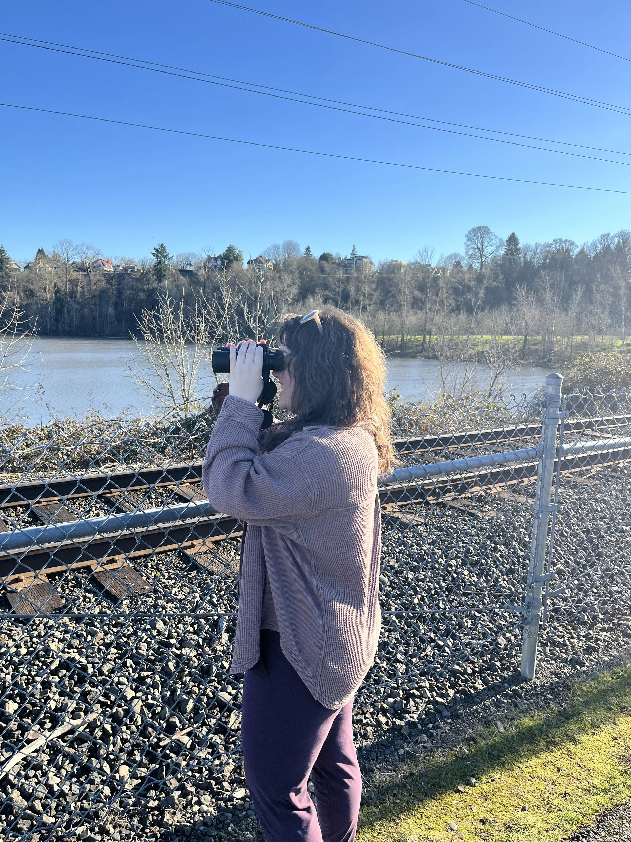 A woman with brown curly hair, wearing a light pink jacket and dark purple pants, is standing outdoors, looking through binoculars across a river with trees visible on the other side under a clear blue sky.
