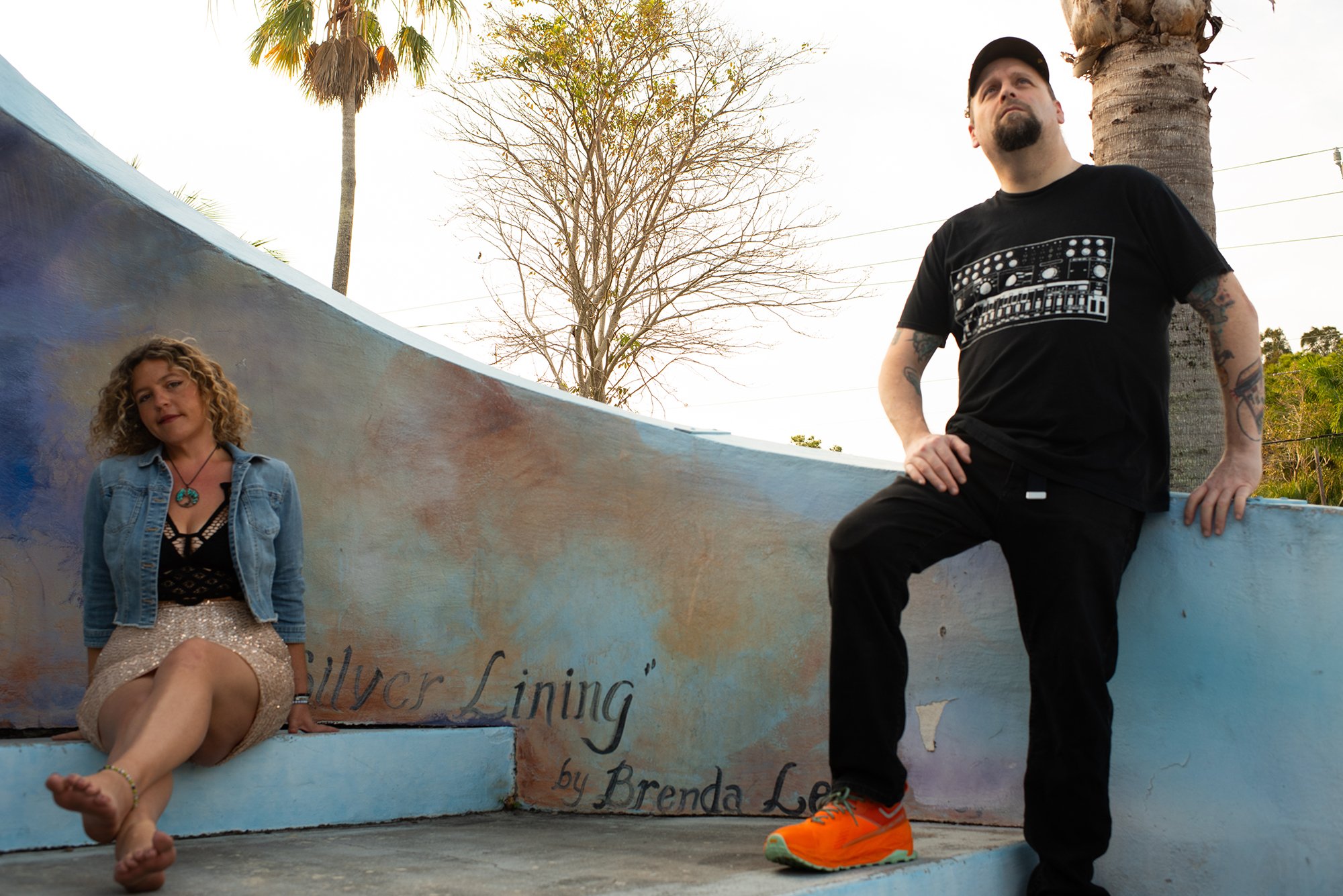 Two people are posing outdoors against a curved wall. The person on the left is sitting, wearing a denim jacket and a sparkly skirt. The person on the right is standing, wearing a black T-shirt and bright orange shoes. Trees and a palm are visible in