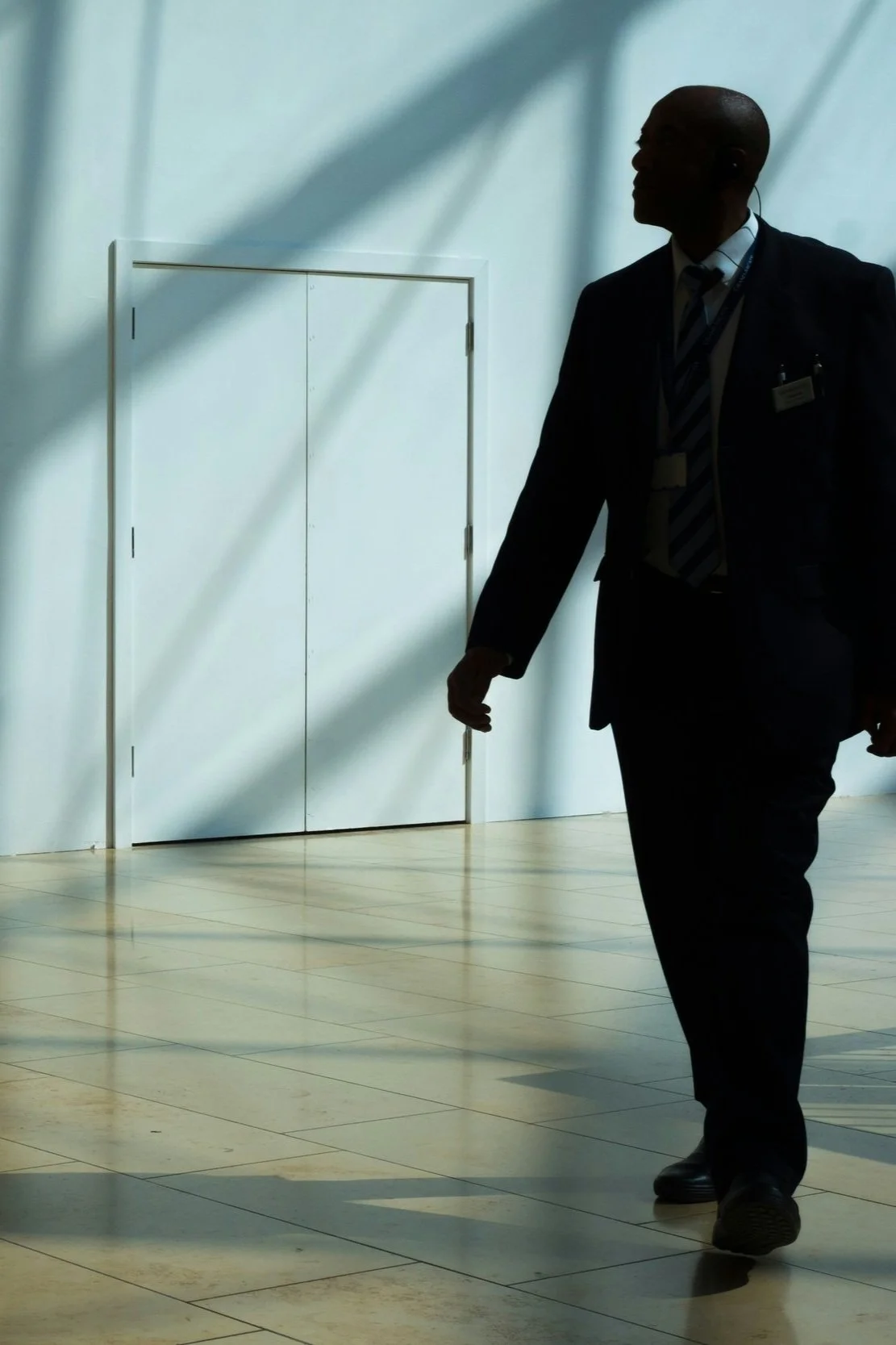 Silhouette of a man in formal attire walking in a sunlit room with a tiled floor and white door.