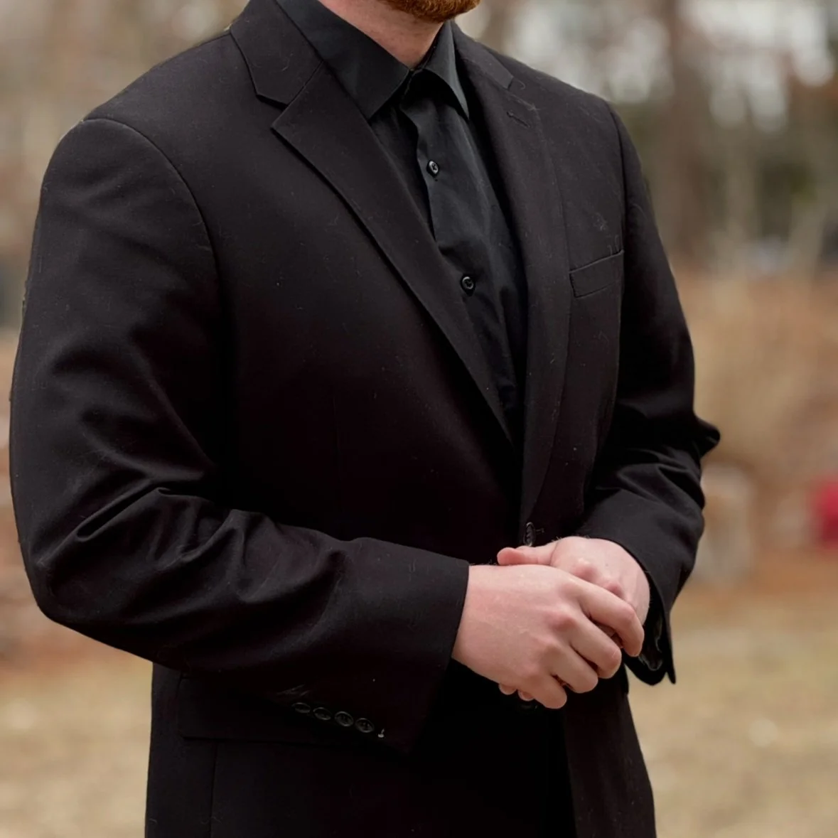 Person in black suit with hands clasped, wearing a black shirt, standing outdoors with blurred background.
