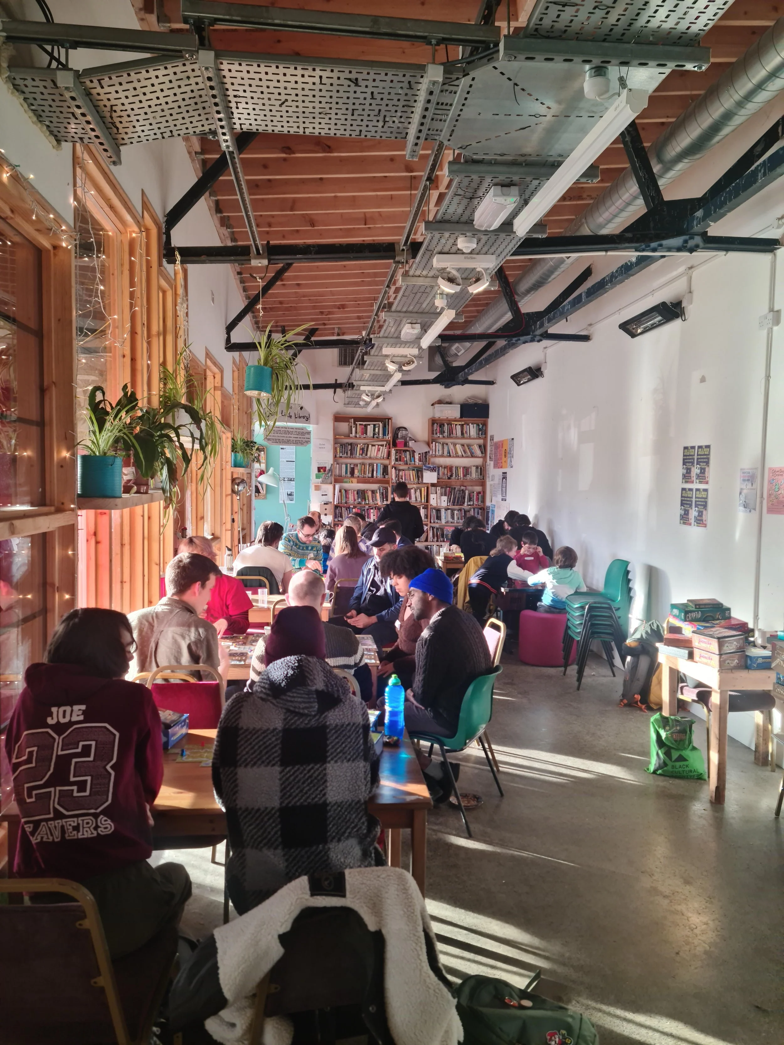 People gathered around tables in a cozy indoor space with wooden walls and bookshelves, playing games and socializing.