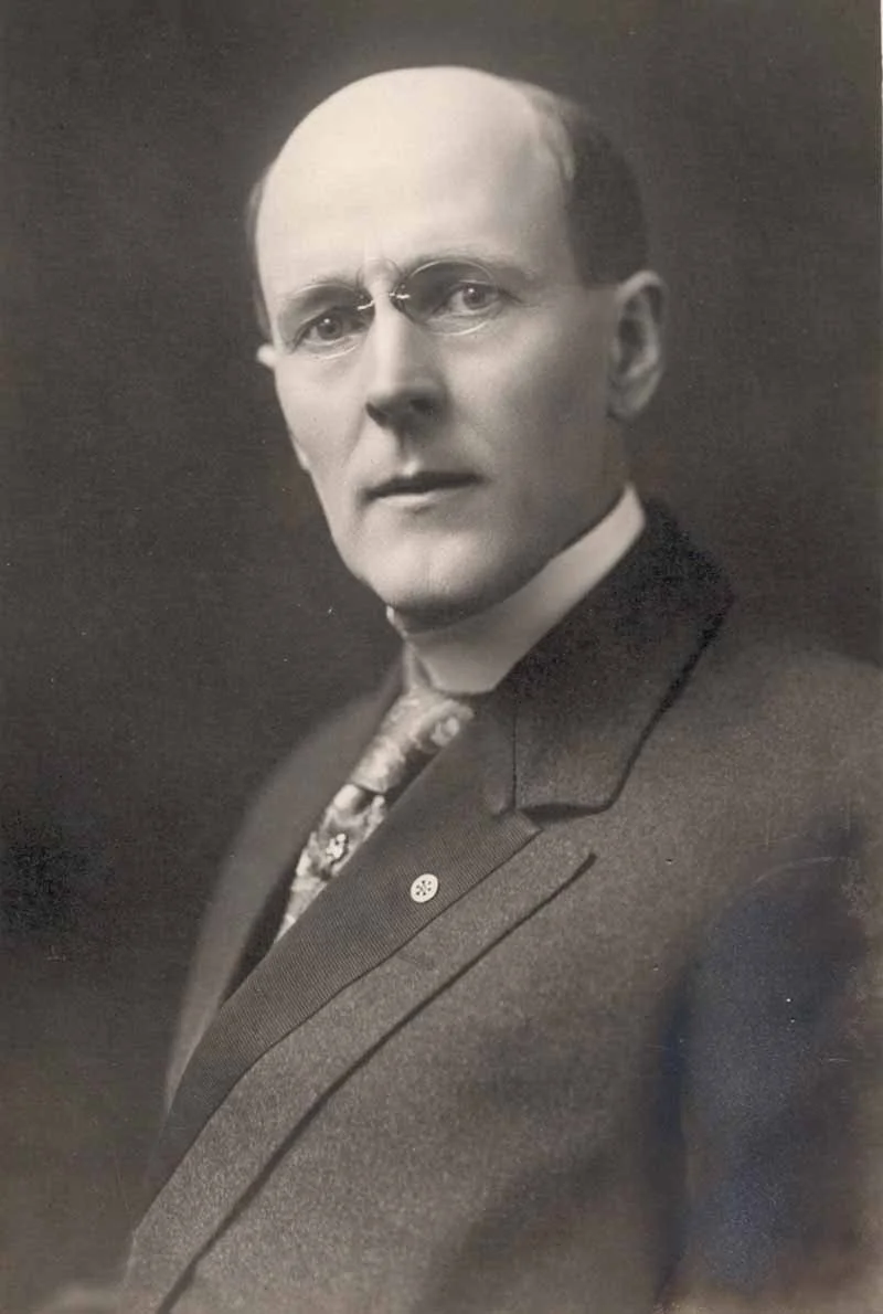 Black and white portrait of a man with glasses, wearing a suit and tie, looking directly at the camera with a serious expression.