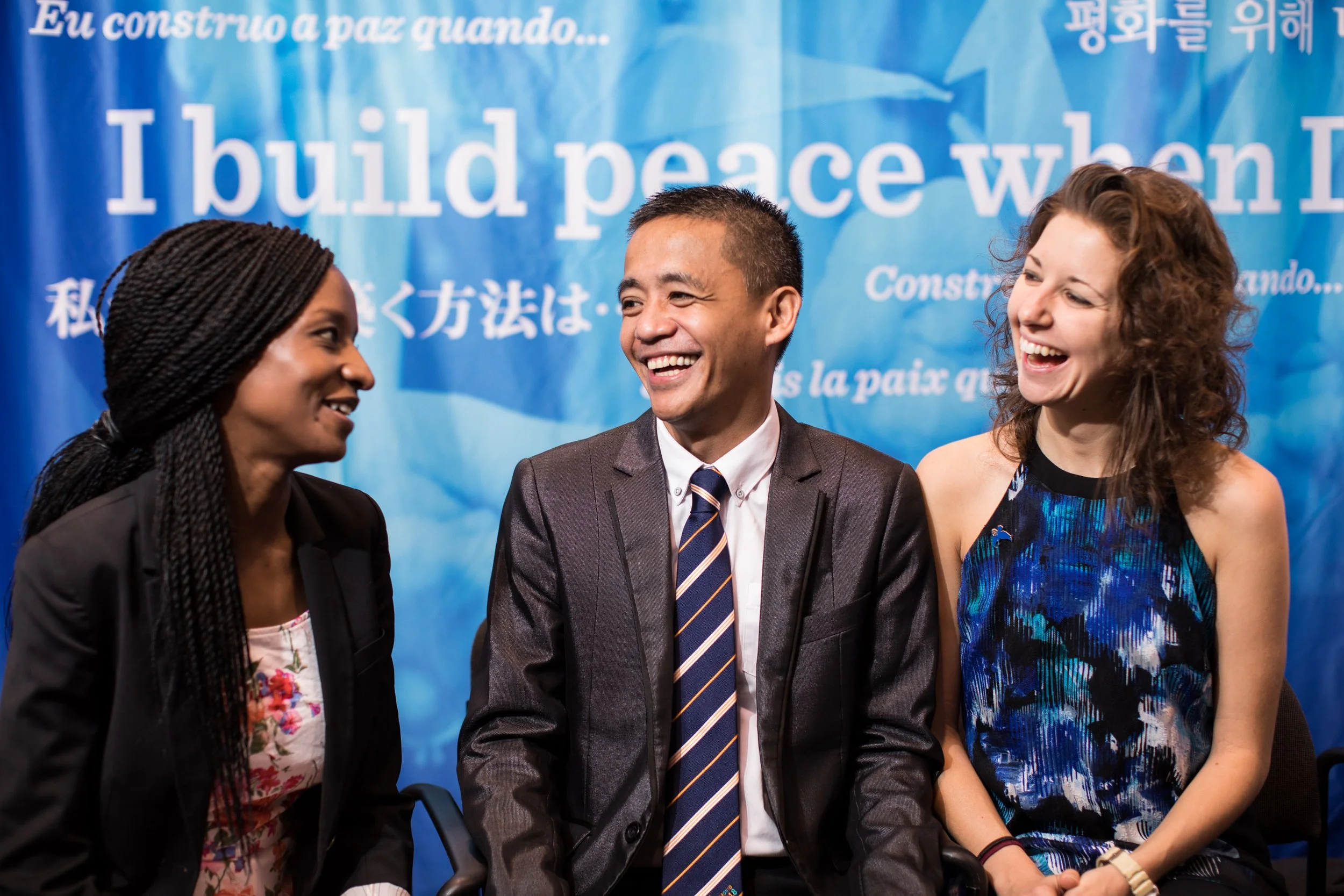 Four diverse people smiling and talking at an event with a blue banner in the background that says 'I build peace when I...'