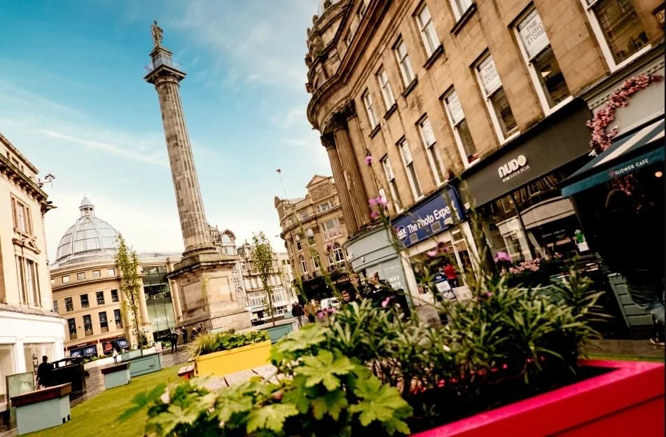 City square with historic buildings, a monument, and storefronts, including a flower bed in the foreground.