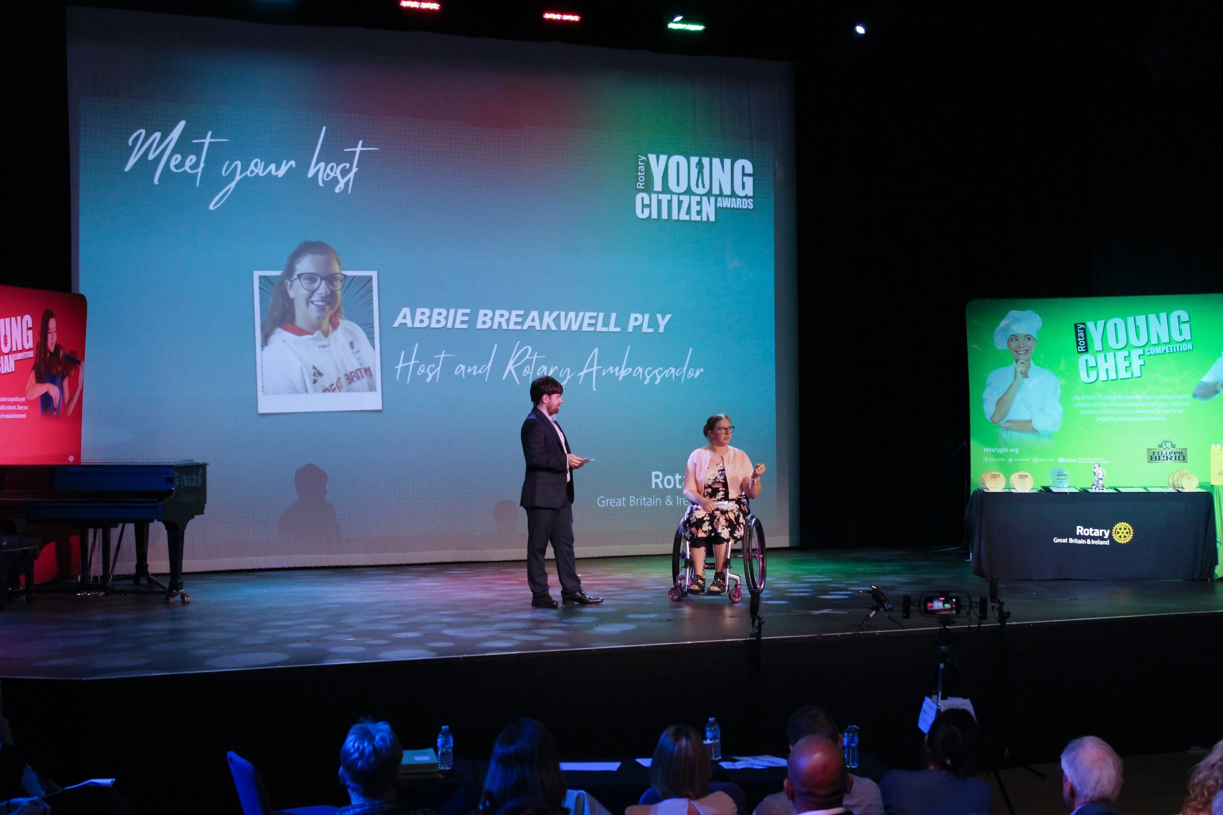 Two people on stage, one standing and one in a wheelchair