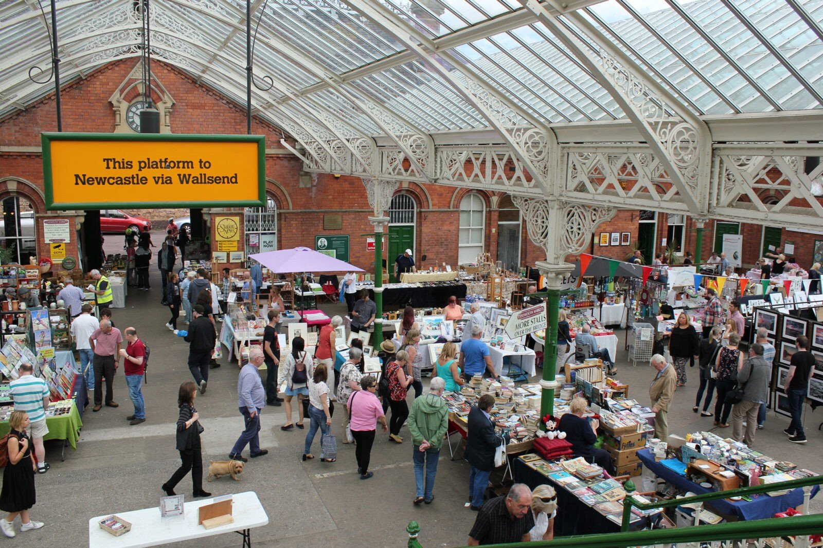 People shopping at market with many tables and booths selling various items under a glass roof with ornate white ironwork and a brick building in the background.