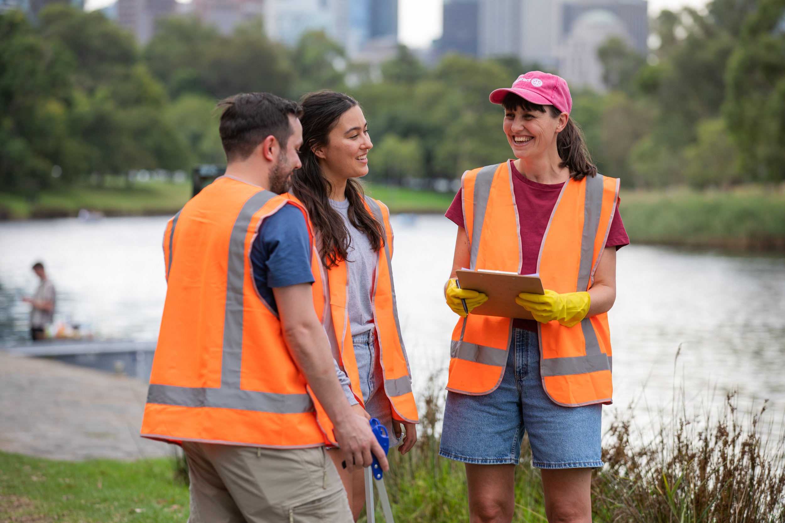 Three women in orange safety vests by a river, smiling and talking, one holding a clipboard, with green trees and cityscape in the background.