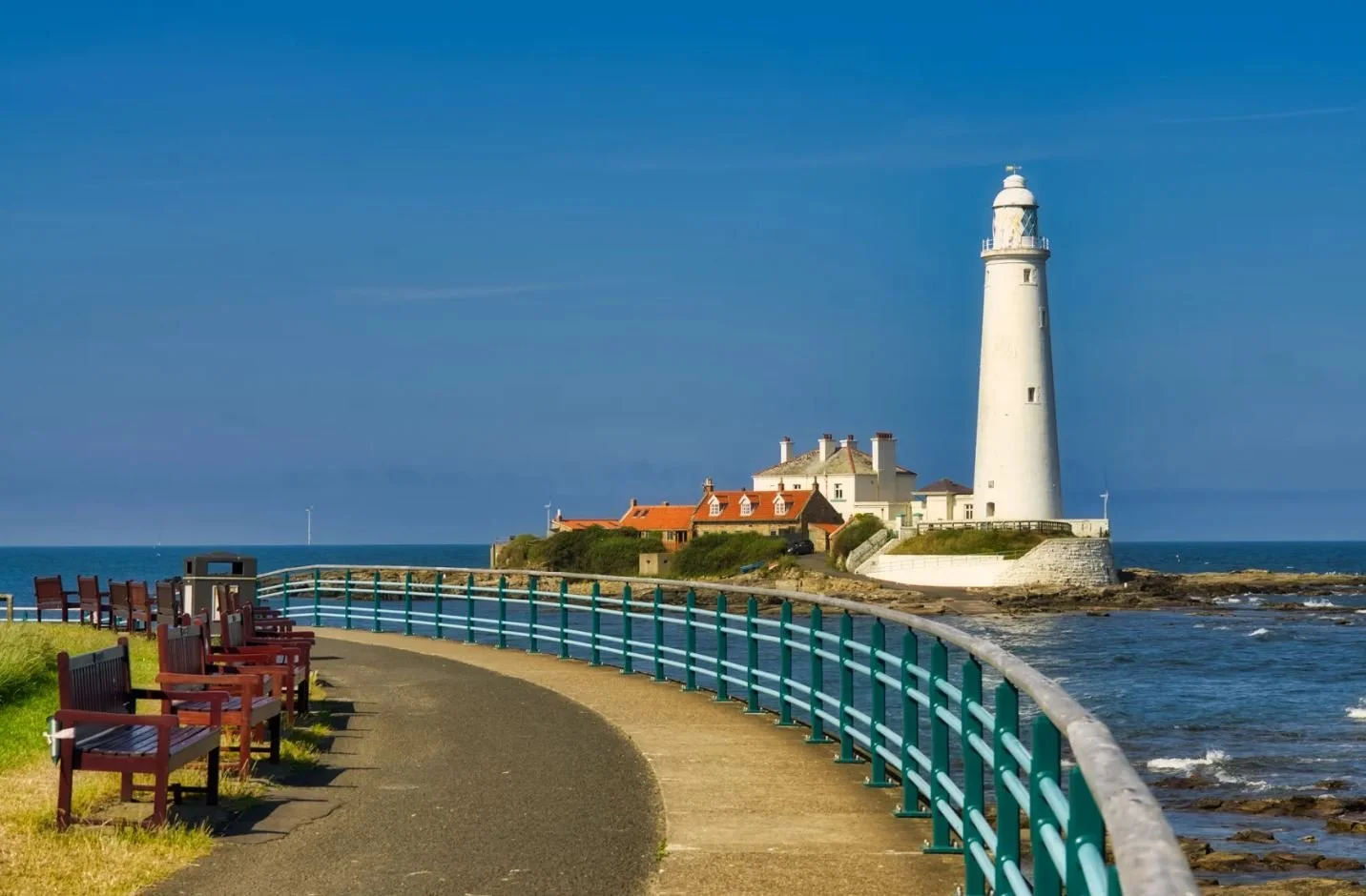 Lighthouse on a rocky coast with a walkway and benches in the foreground, blue sky and ocean in the background.
