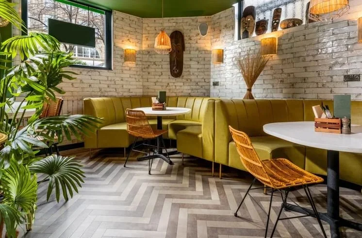 Interior of a cozy cafe with a green ceiling, white brick walls, a large window, a yellow curved booth, round white tables, and wicker chairs. Decor includes hanging lamps, wall-mounted lights, masks on the wall, and potted plants.