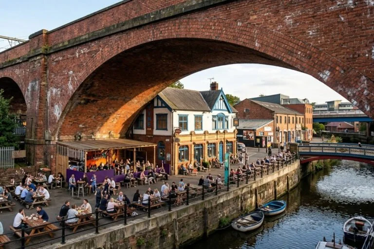 People dining and socializing at outdoor tables along a riverside promenade under a large brick arch.