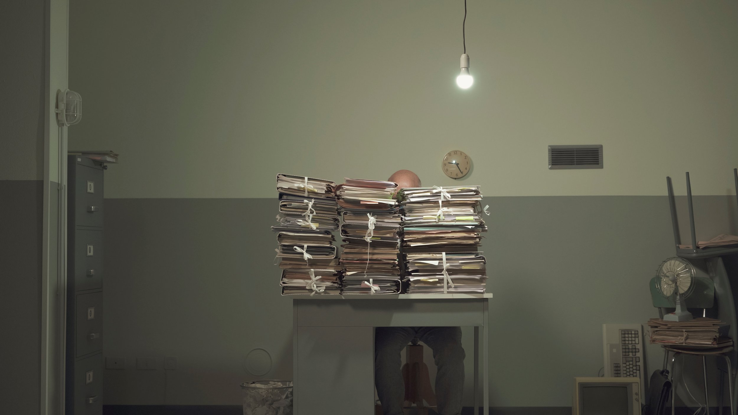 Stacks of case files piled on a desk under a hanging light in a dim office, symbolizing failures in West Virginia’s child welfare system.