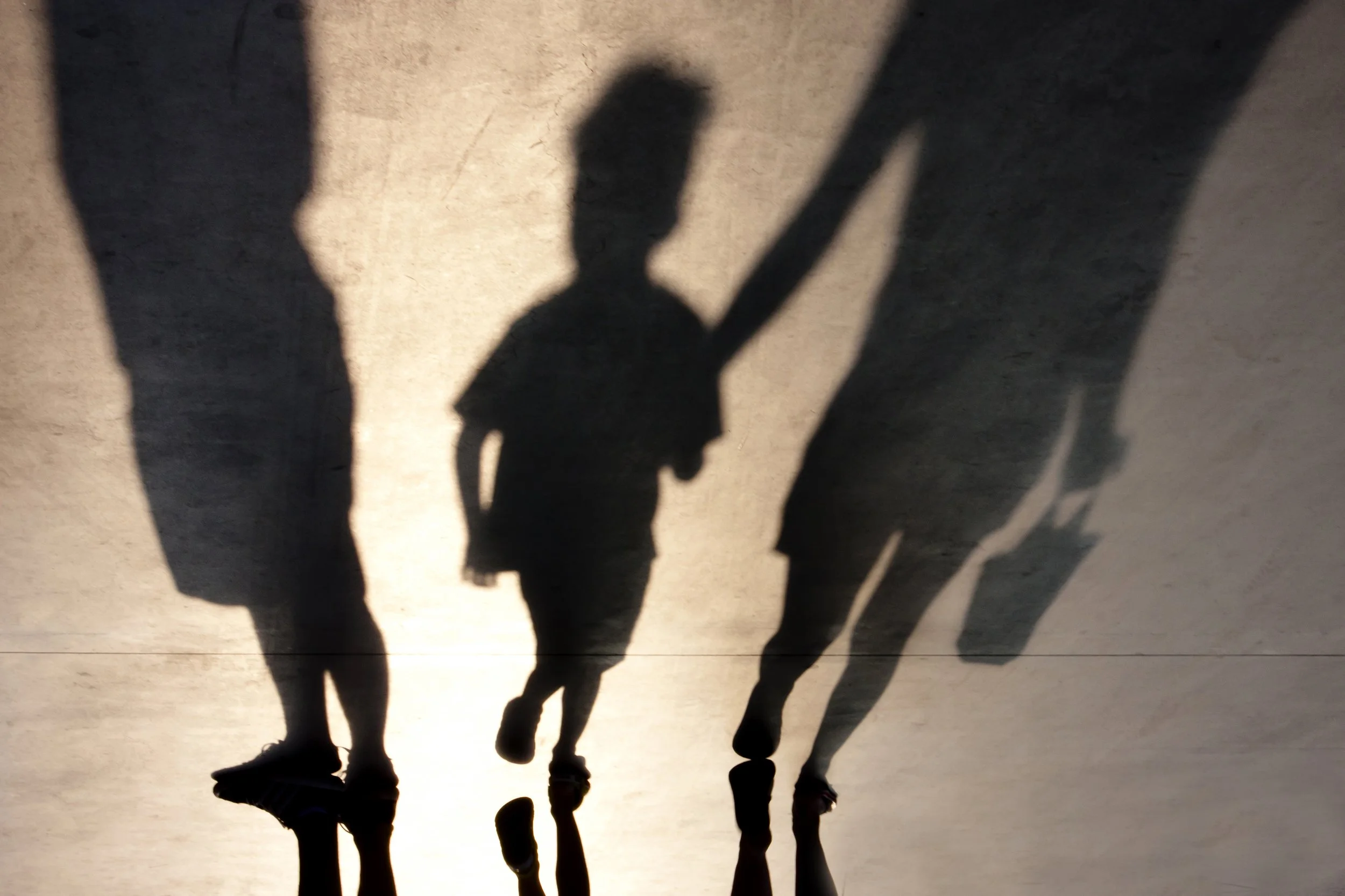 Shadows of a child walking between two adults on a sunlit surface, captured as a silhouette reflection on pavement, evoking themes of separation and state custody.
