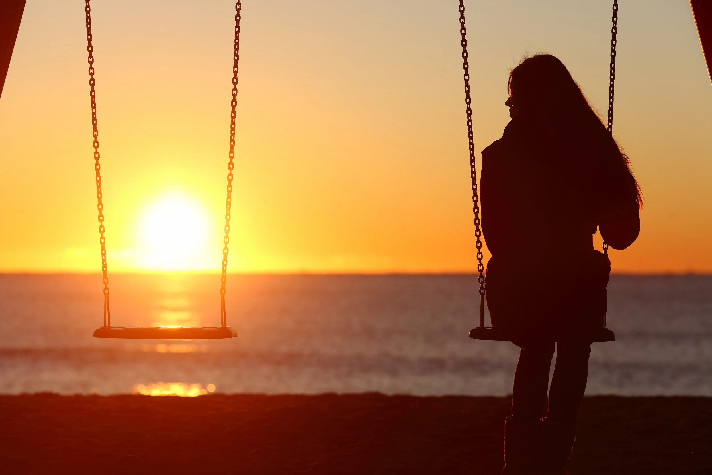 Silhouette of a woman sitting on a swing at sunset beside an empty swing, overlooking the ocean, symbolizing loss and separation in the Abigaile and Natascha case.