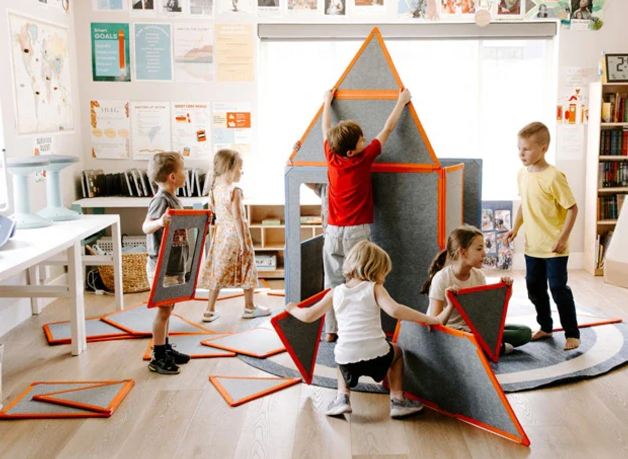 Kids building a soft play structure with large foam panels and orange frames in a classroom.
