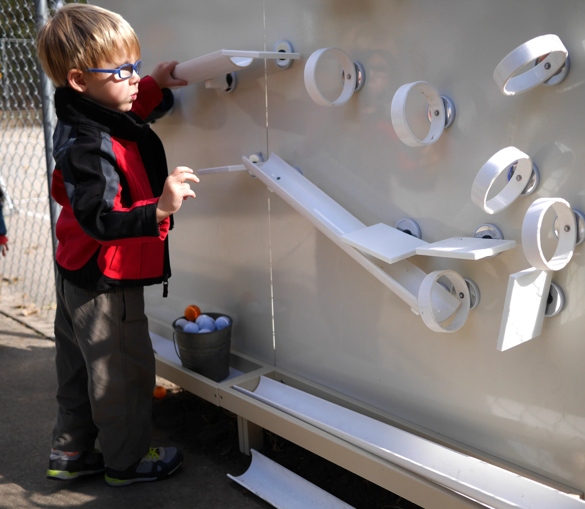A young boy with glasses playing a game of skee-ball at an outdoor arcade. He is wearing a red and black jacket, gray pants, and black shoes, and is aiming to roll a ball up the inclined lane.