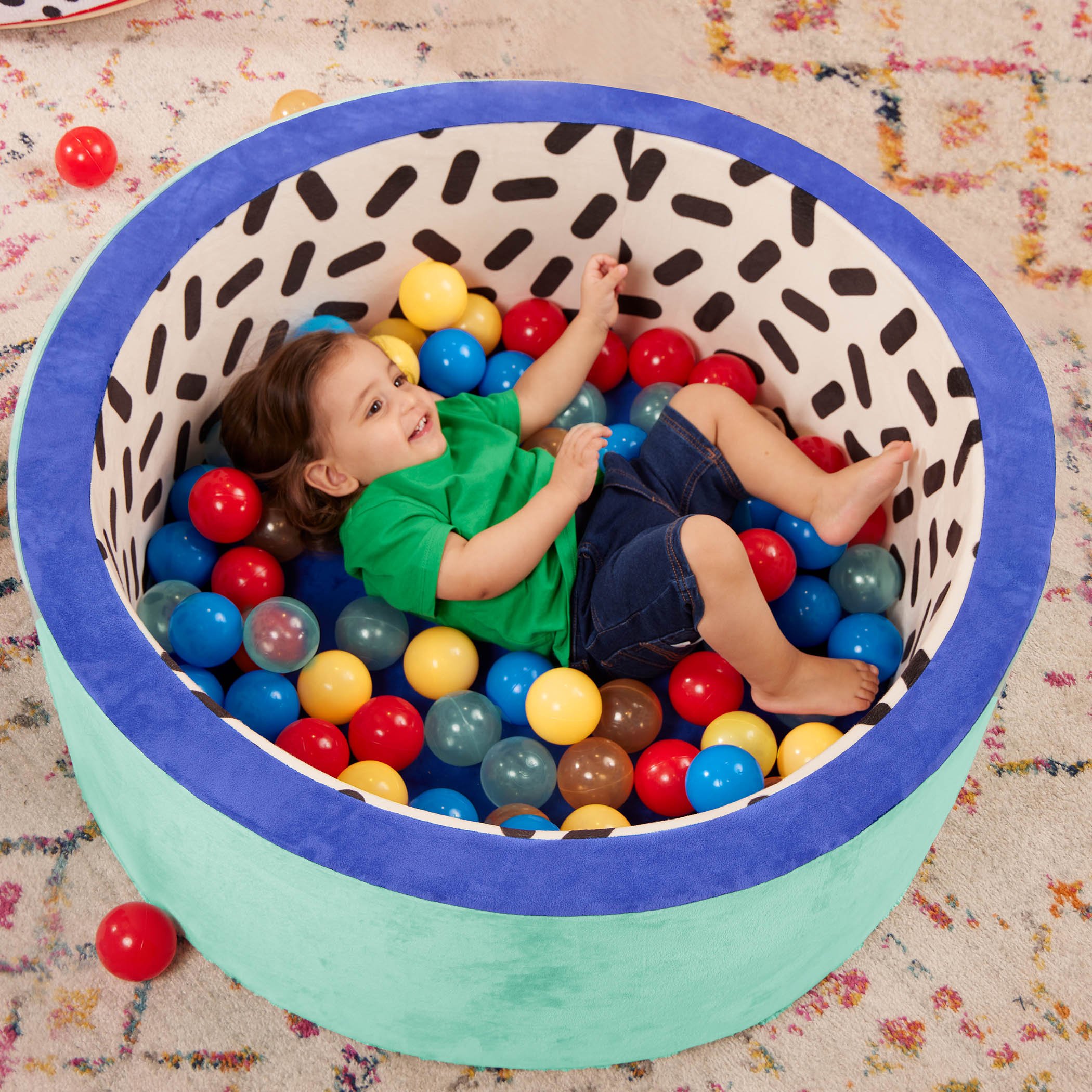 A young child with brown hair wearing a green shirt and denim shorts lying in a ball pit filled with colorful plastic balls on a carpeted floor.