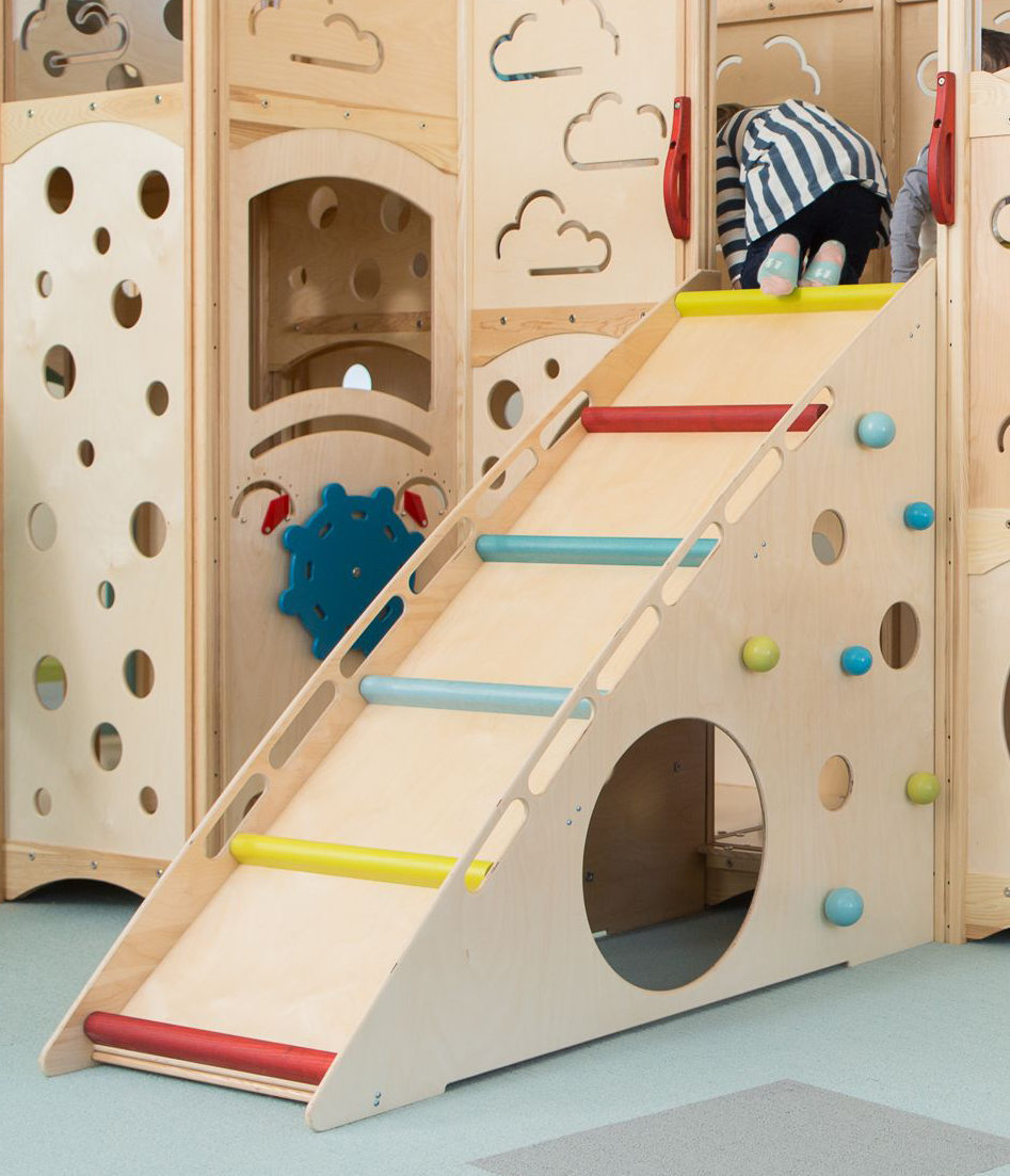 Children playing on a wooden indoor playground with a slide, climbing elements, and various cutouts, in a room with light-colored flooring.