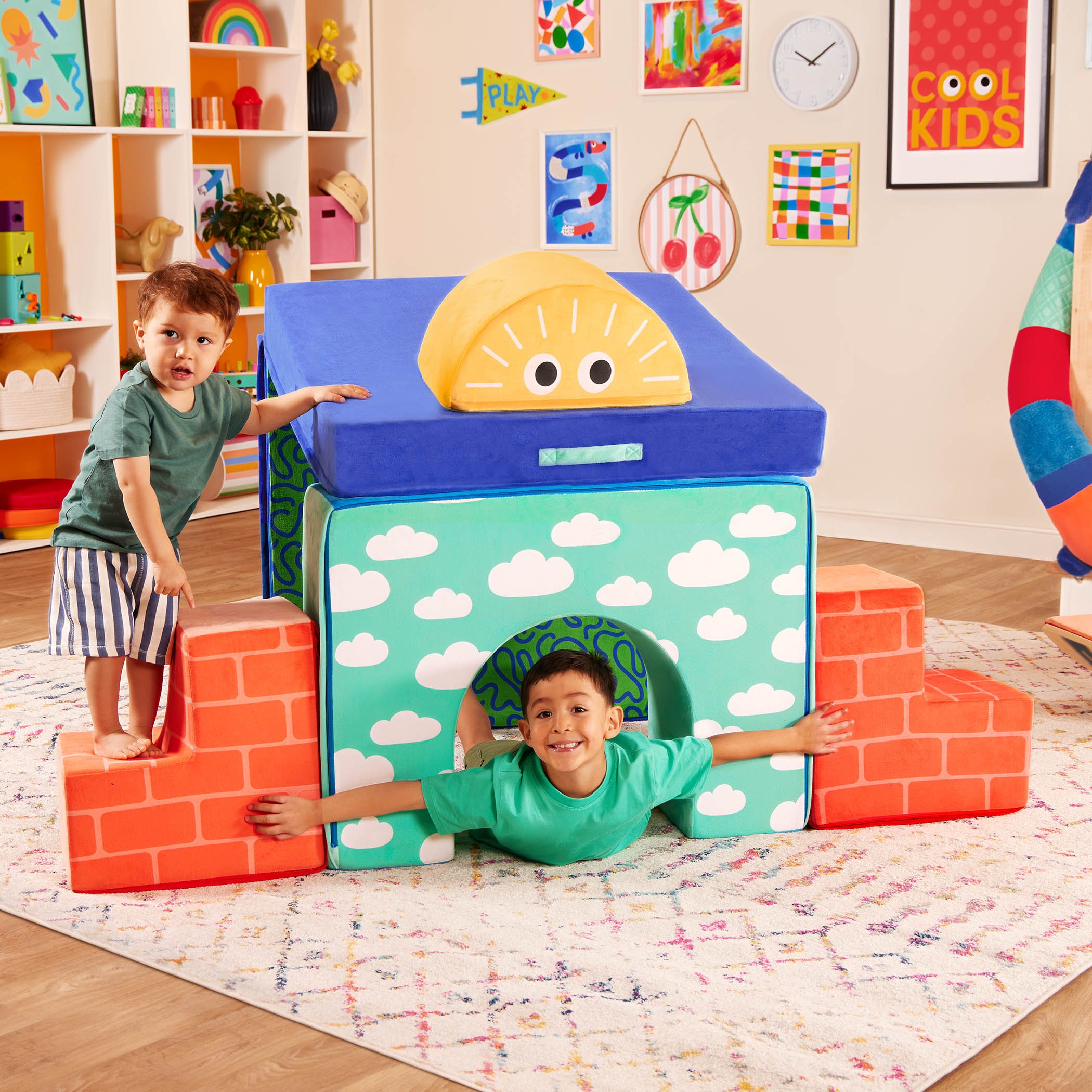 Two young boys playing inside a colorful, soft fabric playhouse that looks like a toy brick oven in a cheerful playroom decorated with colorful art and toys.