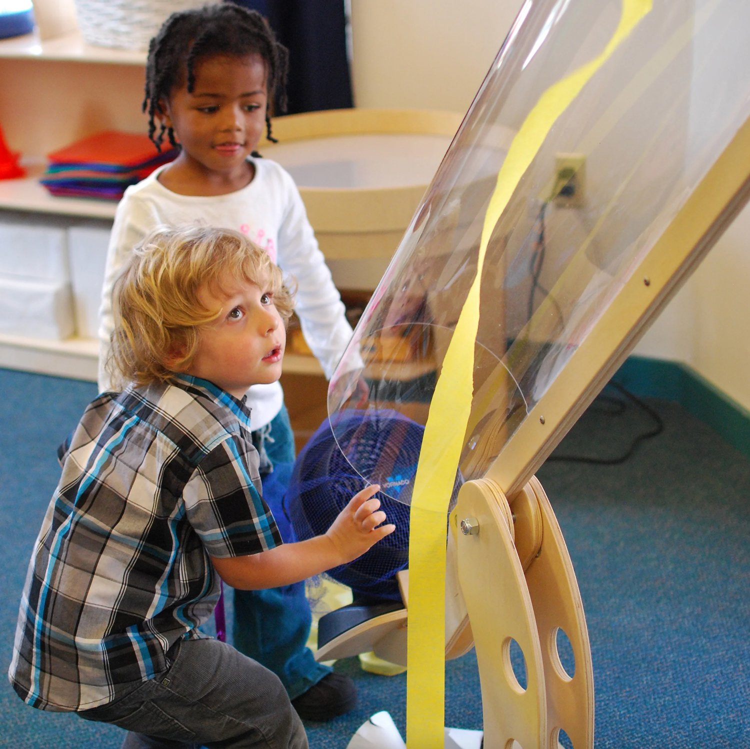 Two children, a girl and a boy, are looking at a clear plastic exhibit with yellow tape on it, in an indoor setting with shelves and colorful objects in the background.