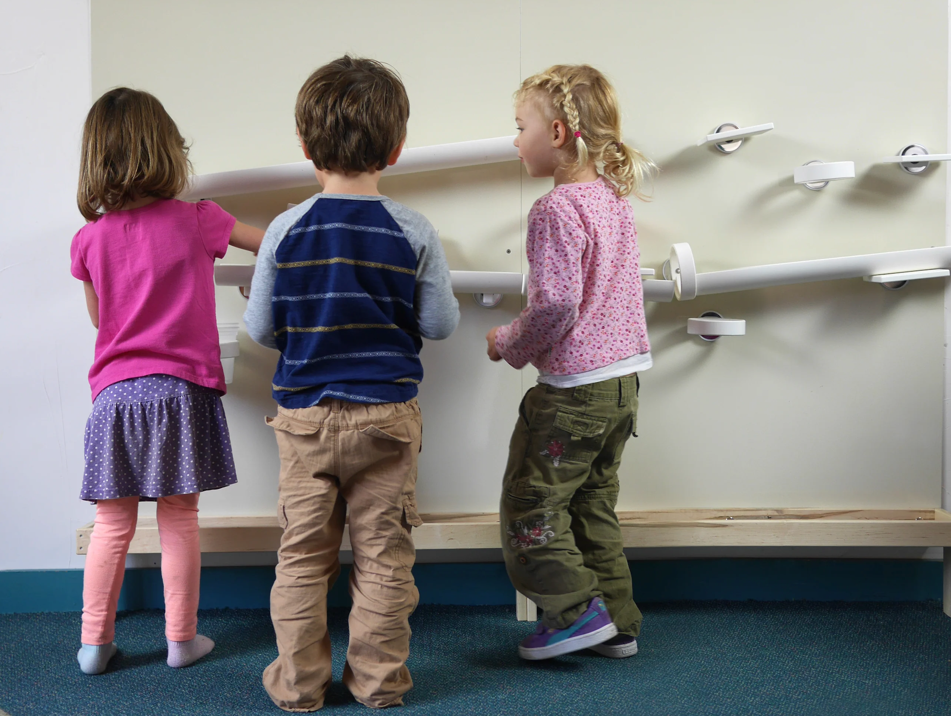 Three young children, two girls and one boy, viewing from the back, stand in front of a large white wall with multiple white horizontal bars at different heights.