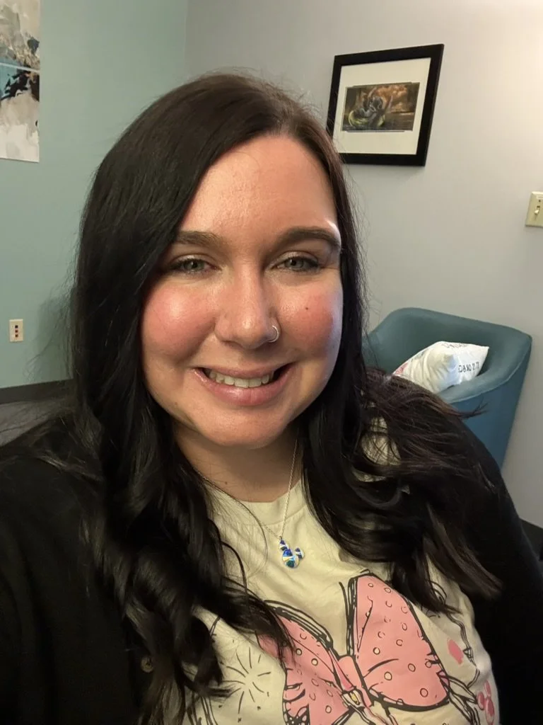 A woman with long dark hair and a nose ring smiling, wearing a butterfly graphic t-shirt and a blue necklace, in a room with light green walls and framed artwork.
