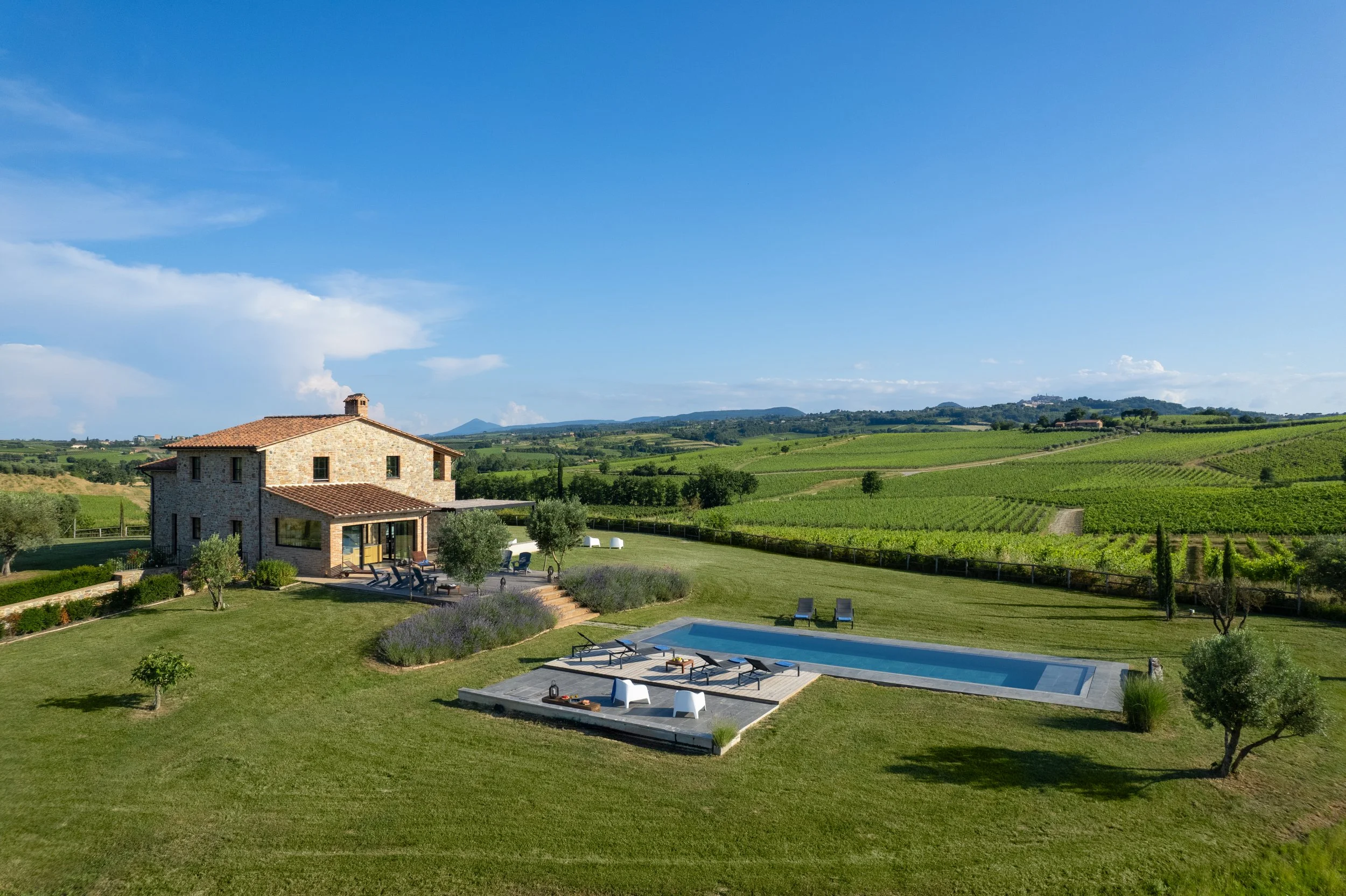 Rustic stone house with a tiled roof in a lush green landscape, with a swimming pool and outdoor seating area, rolling hills and vineyards in the background under a blue sky.