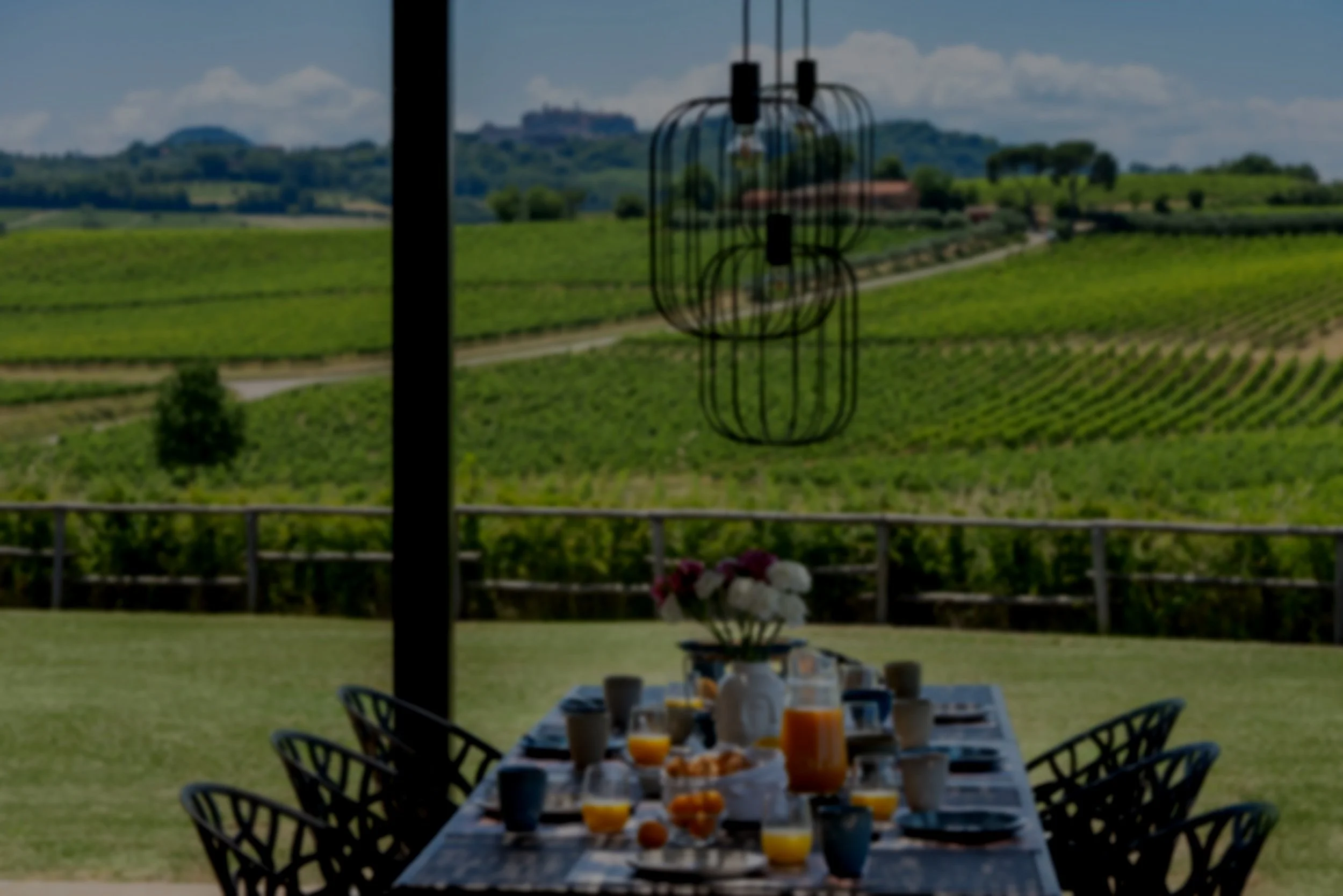 An outdoor dining table set for breakfast with orange juice, coffee, flowers, and breakfast items, overlooking a vineyard with rolling hills and a castle in the background.