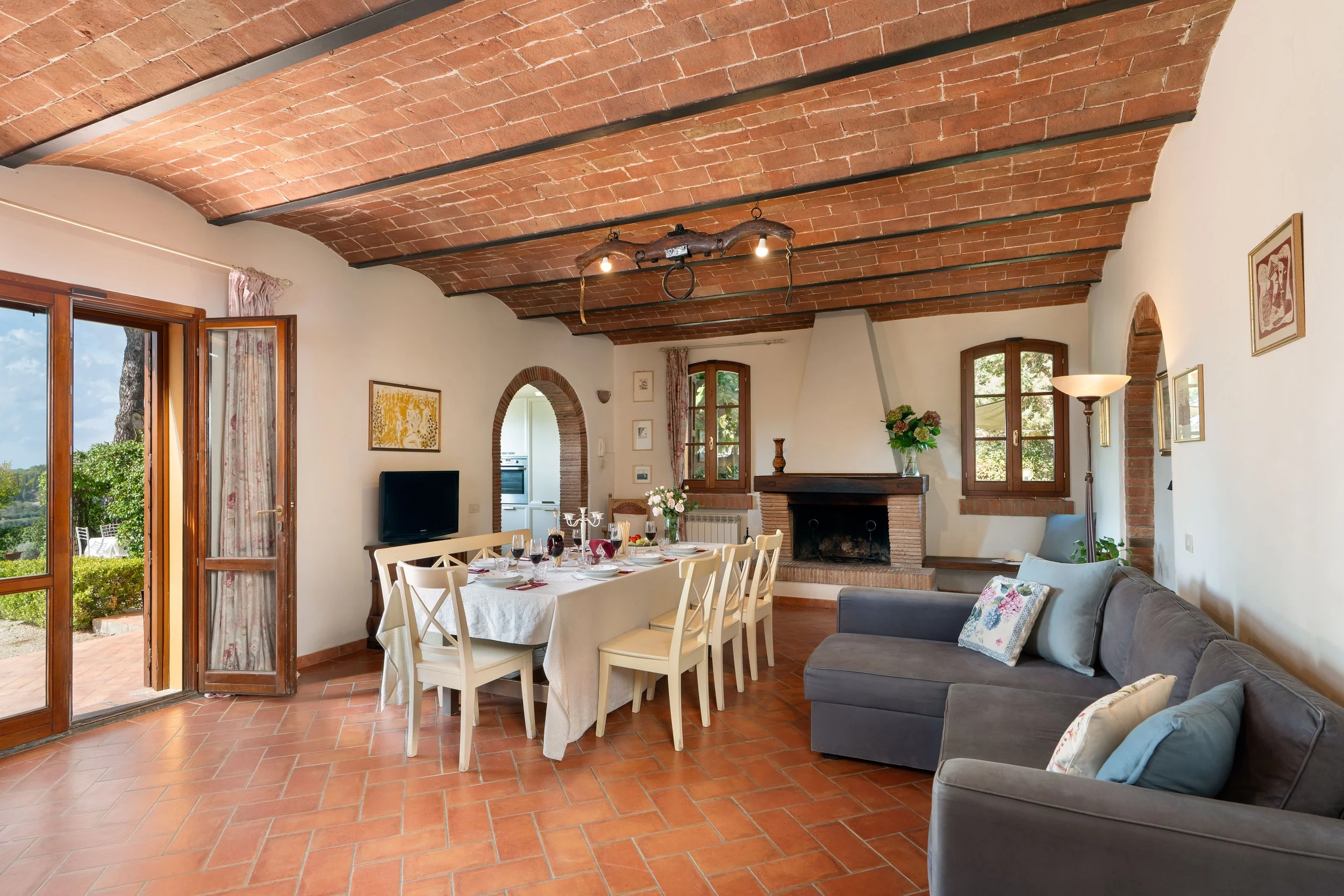Living room with terracotta tile floor, brick arched ceiling, and white walls. Features a dining table set with dishes and wine glasses, a gray sofa with pillows, a fireplace, three wooden framed windows, and a door leading outside to a garden.