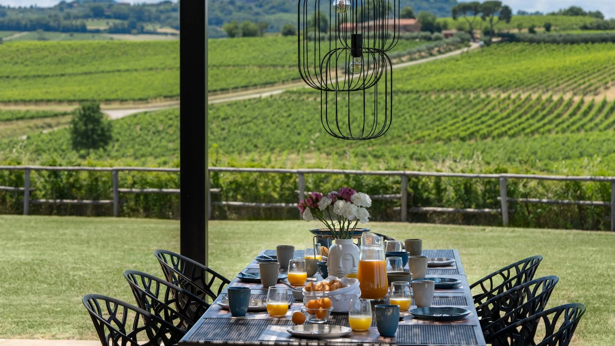 A table set for breakfast outdoors overlooking a lush green vineyard, with a vase of pink and white flowers in the center, glasses of orange juice, cups, plates, and croissants.