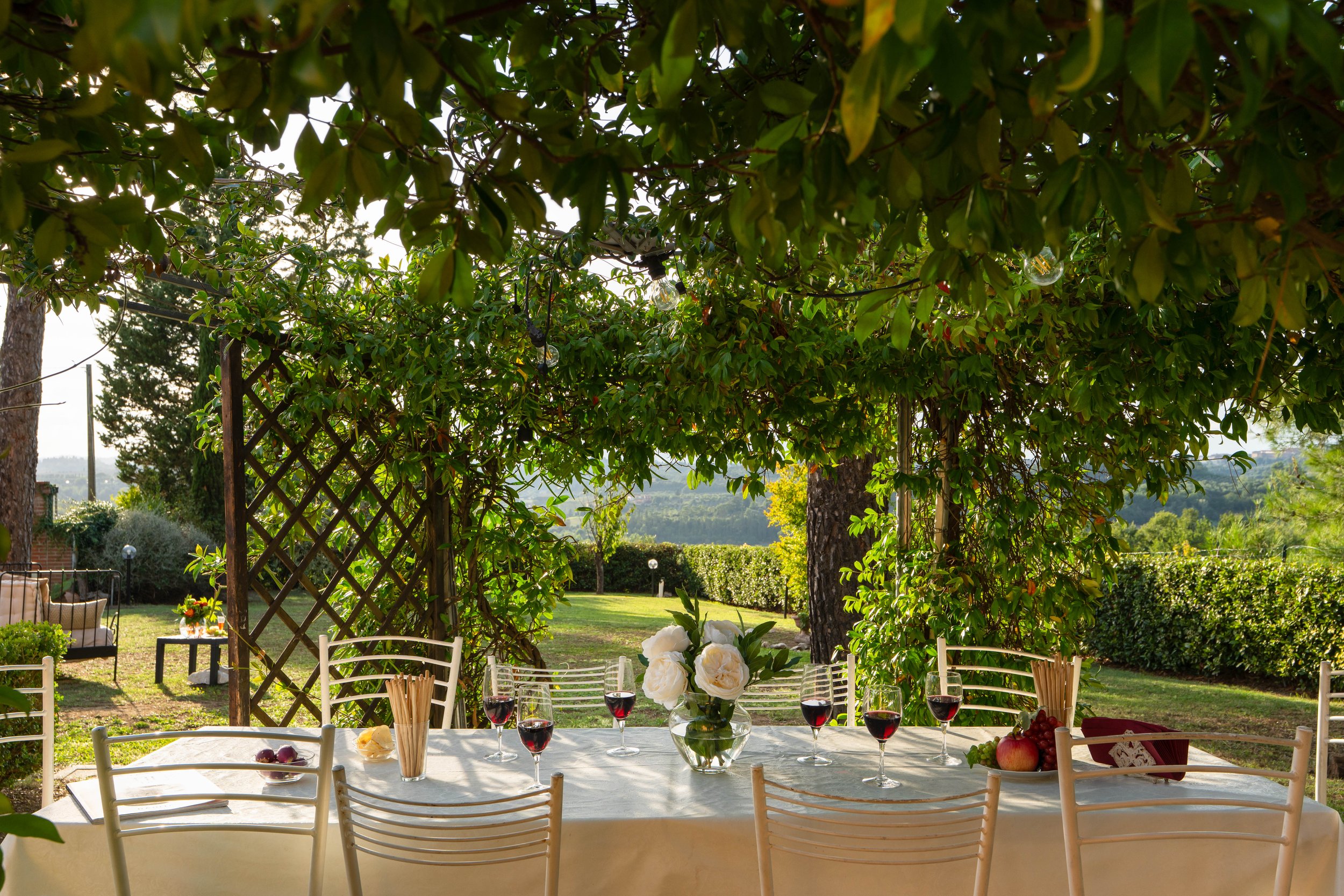 Outdoor dining table set for a meal with wine glasses, a flower vase, and a bowl of fruit, under a green leafy tree in a garden with lush grass and hedges.