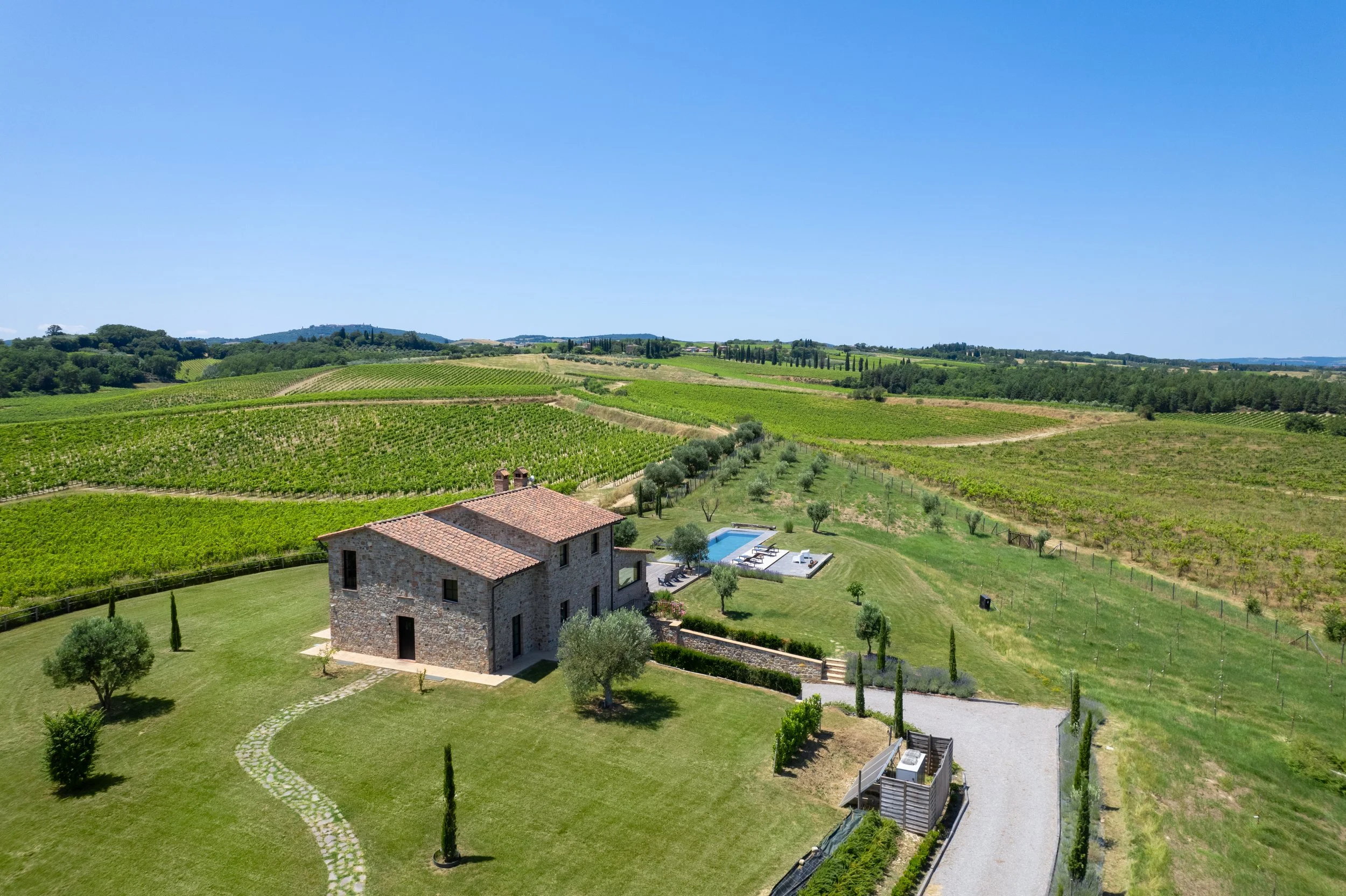 A rustic stone house with a tiled roof, surrounded by a well-maintained lawn with small trees and cypress, is near a swimming pool with lounge chairs. The property is set in a rural landscape of rolling vineyards, fields, and hills under a clear blue