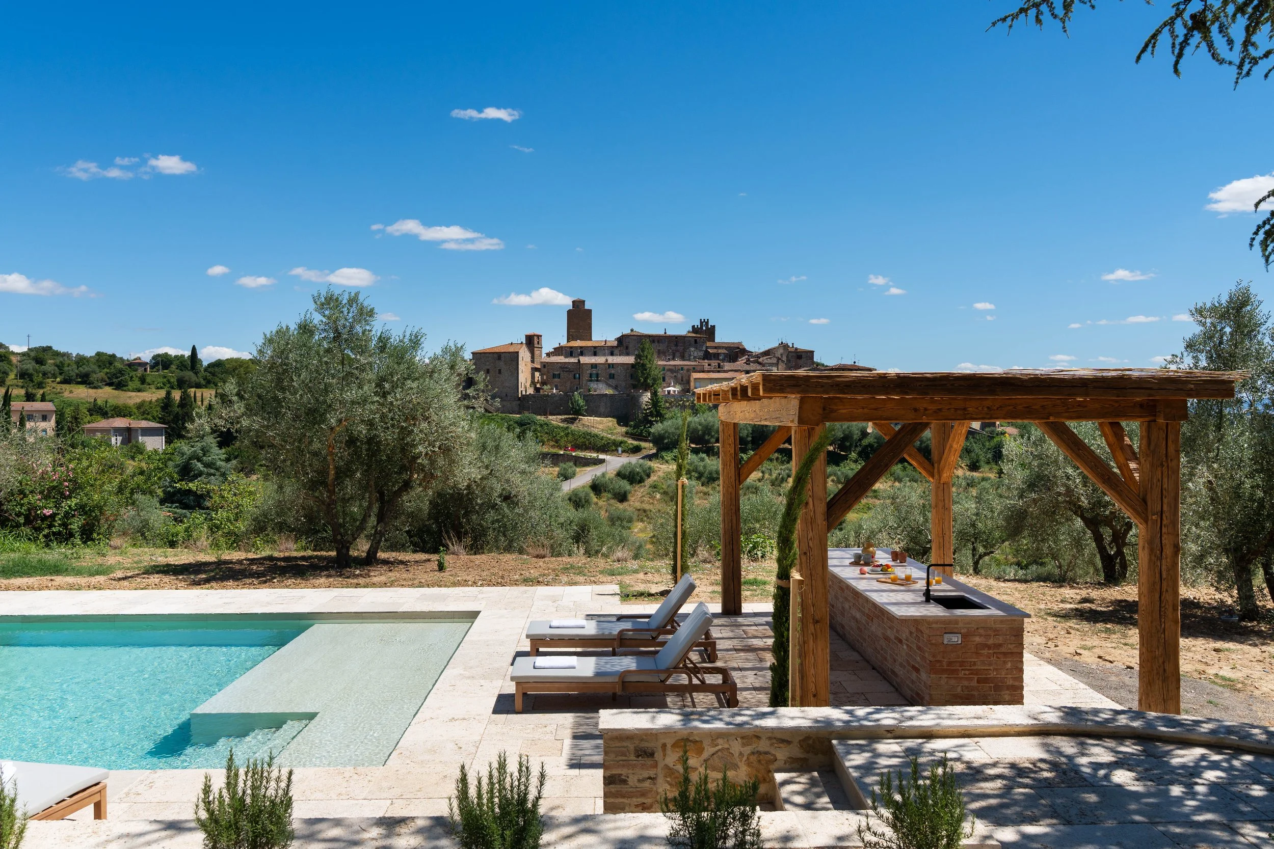 Outdoor poolside area with a swimming pool, lounge chairs, and a wooden outdoor kitchen with a roof, overlooking a scenic hillside village under a clear blue sky.