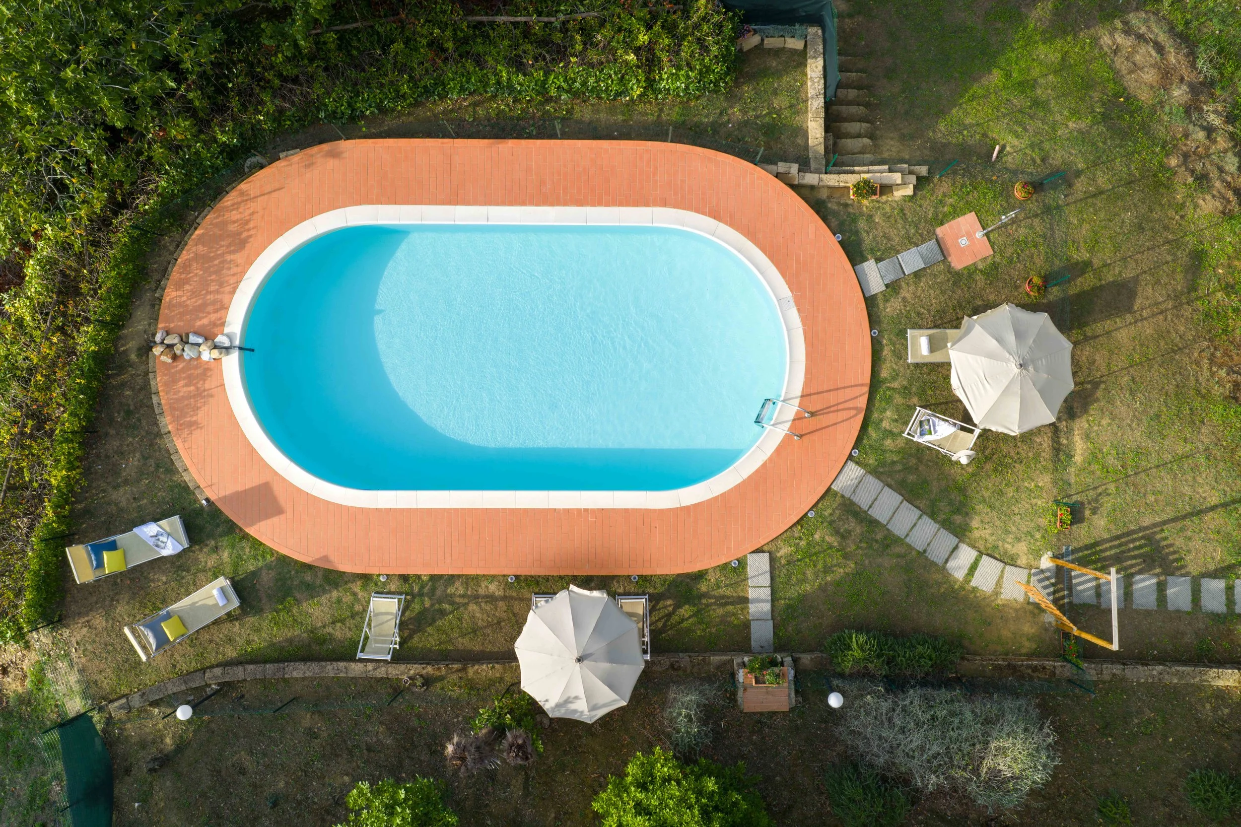 A backyard with an oval swimming pool, surrounded by a red-brick patio, green grass, trees, and outdoor furniture including umbrellas, lounge chairs, and a sandbox.