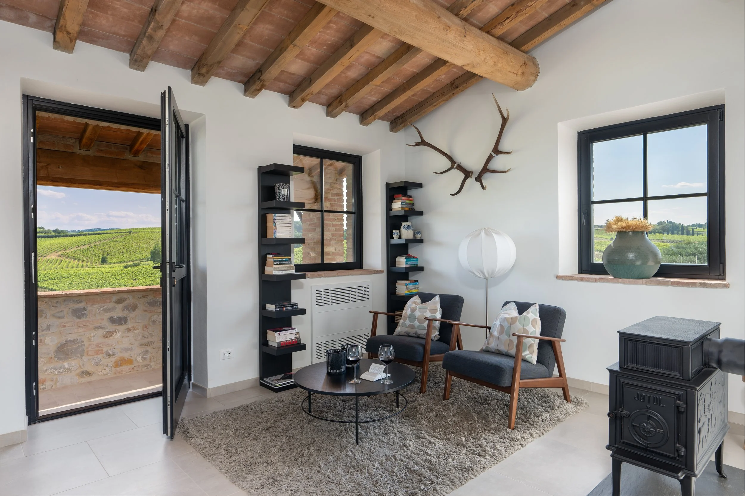 Cozy living room with two armchairs, a round coffee table, bookshelves, and a wood stove, with a window showing a scenic view of vineyards.